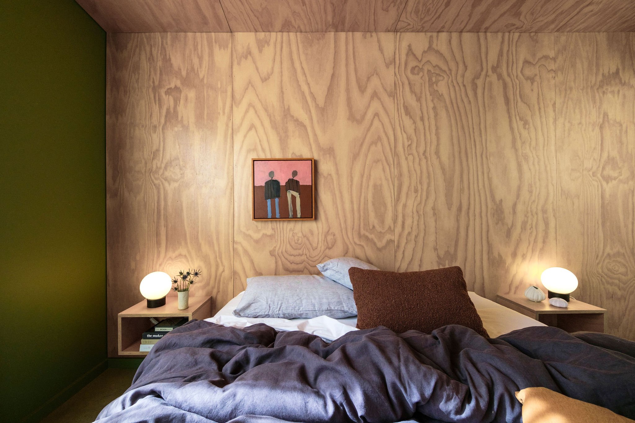 A bedroom with a wooden panel wall, green accent wall, and two bedside tables with spherical lamps, a vase with flowers, and books. A bed with pillows and dark bedding is in the foreground, with a framed painting of two people hanging on the wall above the bed.