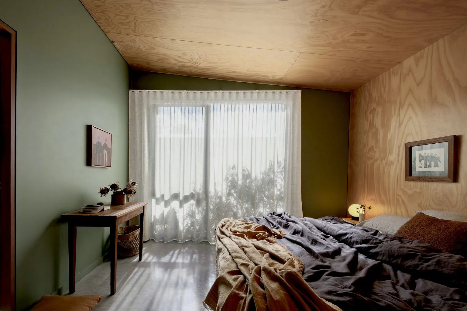 A cozy bedroom with a wooden ceiling and walls, a large window with sheer curtains, a bed with rumpled blankets, a nightstand with a small lamp, and framed artwork on the walls.