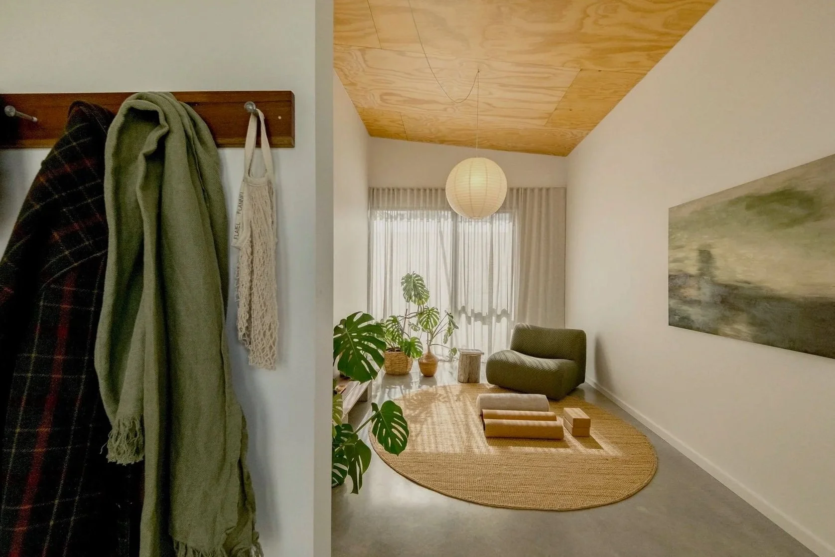 A cozy living room with light-colored walls, a wooden ceiling, and a round beige rug. There are potted plants, a green armchair, and a large abstract painting on the wall. A paper lantern hangs from the ceiling, and sheer curtains cover the window, allowing natural light to fill the space.