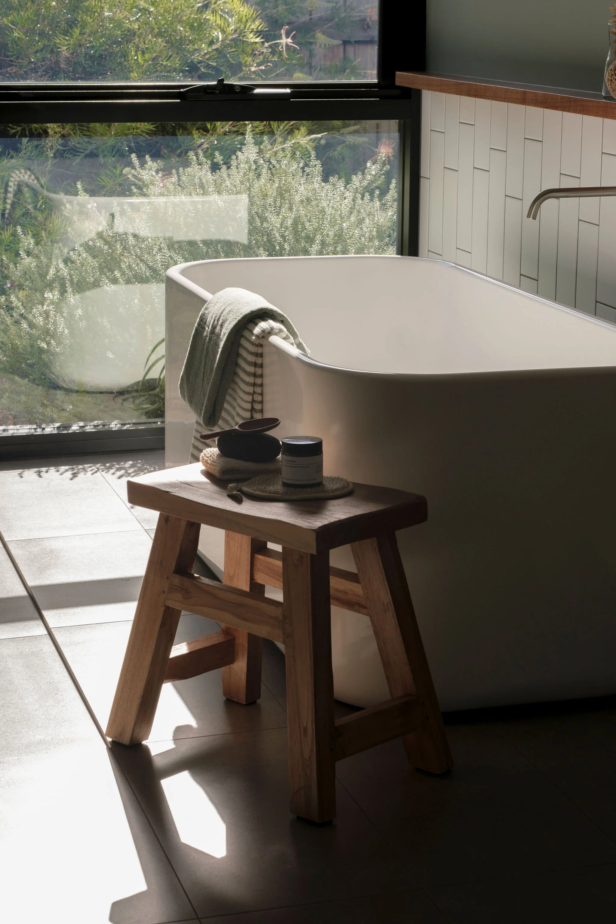 A modern bathtub with a striped towel hanging on its side, placed in a room with natural sunlight coming through a large window. The window looks out onto greenery outside. A small wooden table with bath products and a towel is next to the tub.