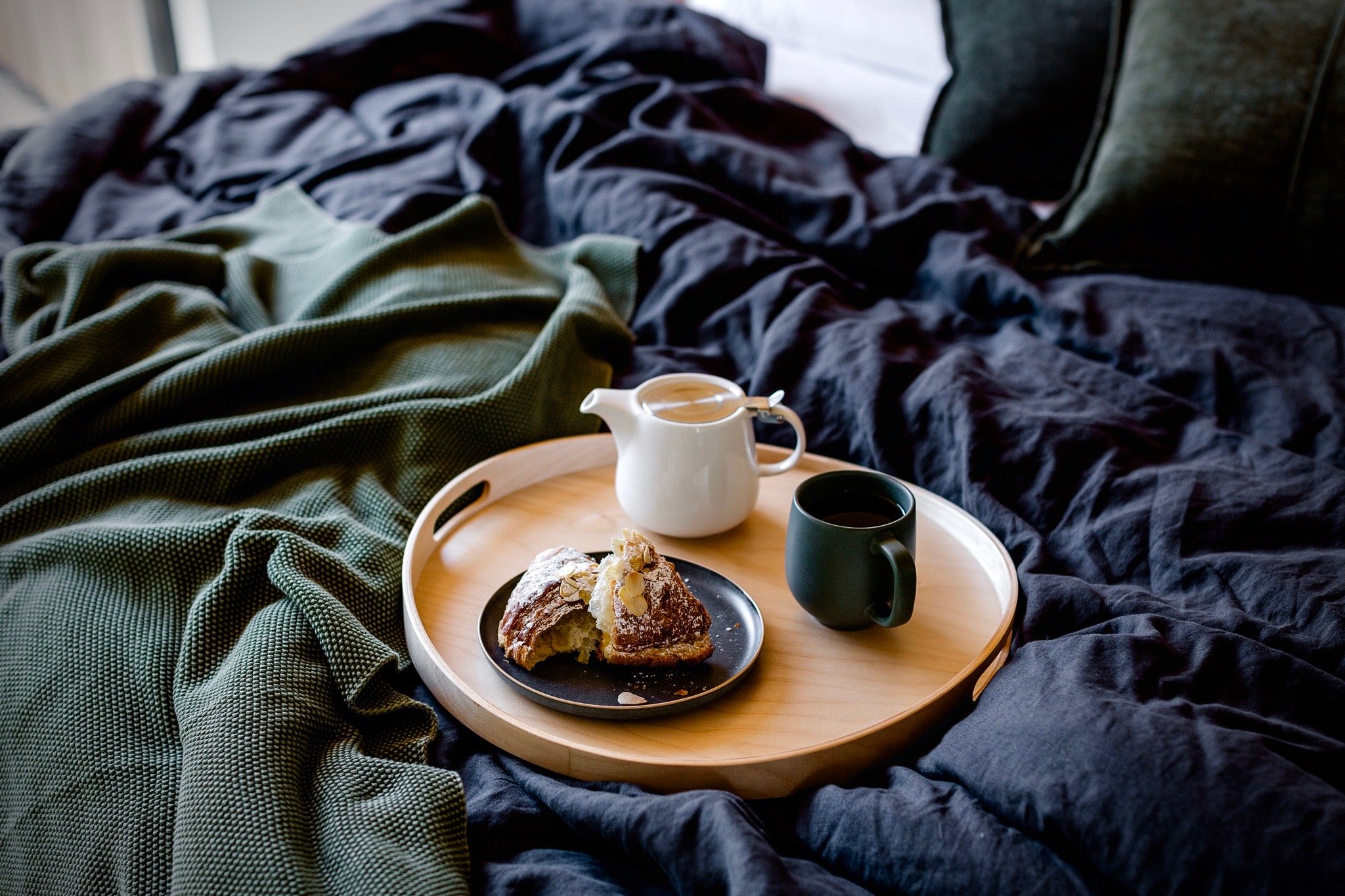 A tray with coffee, a piece of cake topped with almonds, and a pitcher of milk on a bed with dark bedding and green blanket.