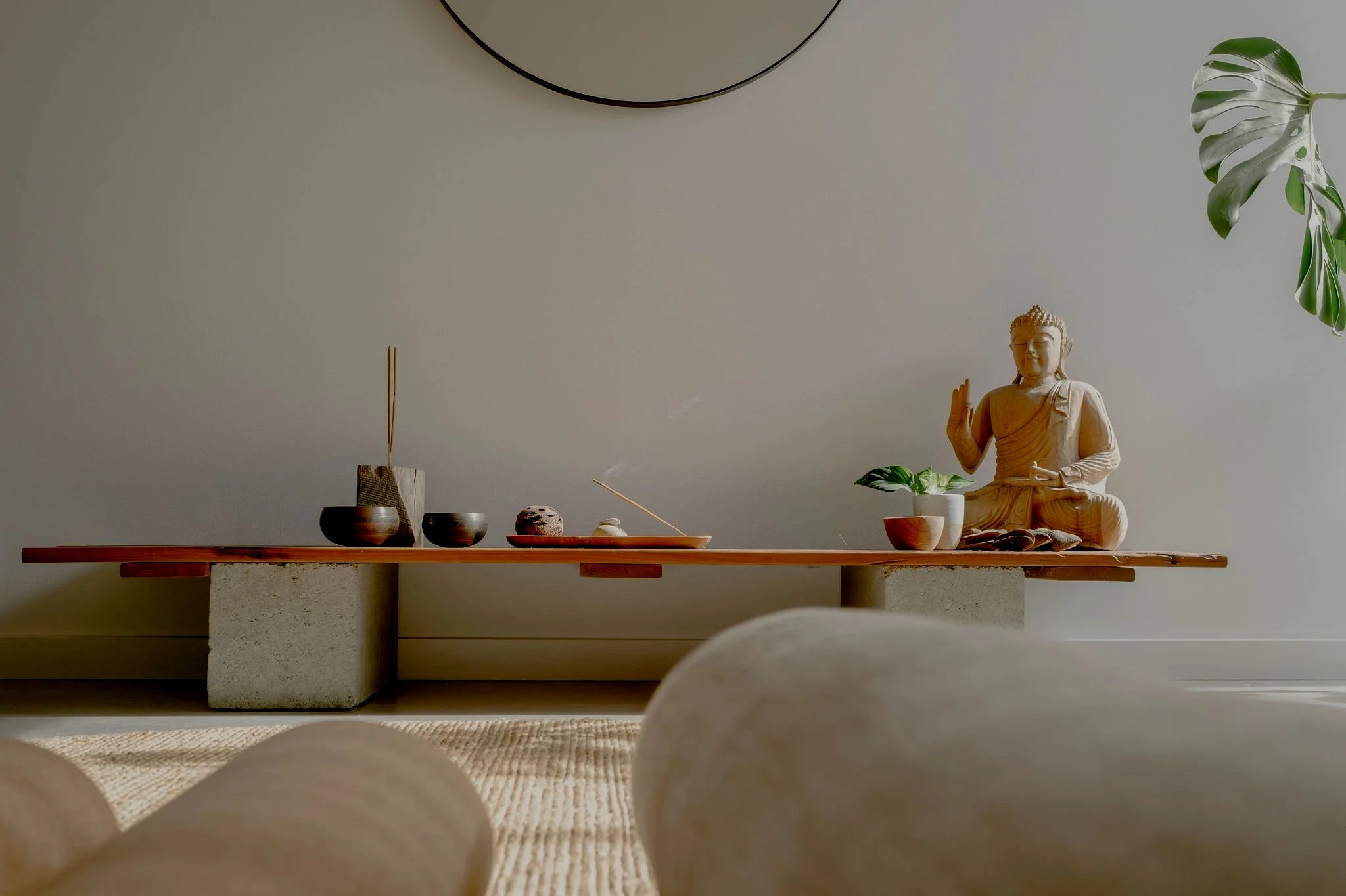 A minimalist altar features a wooden table on concrete supports with various objects including a small Buddha statue, incense, stones, and plants against a plain wall with a large circular mirror and a green plant with split leaves.