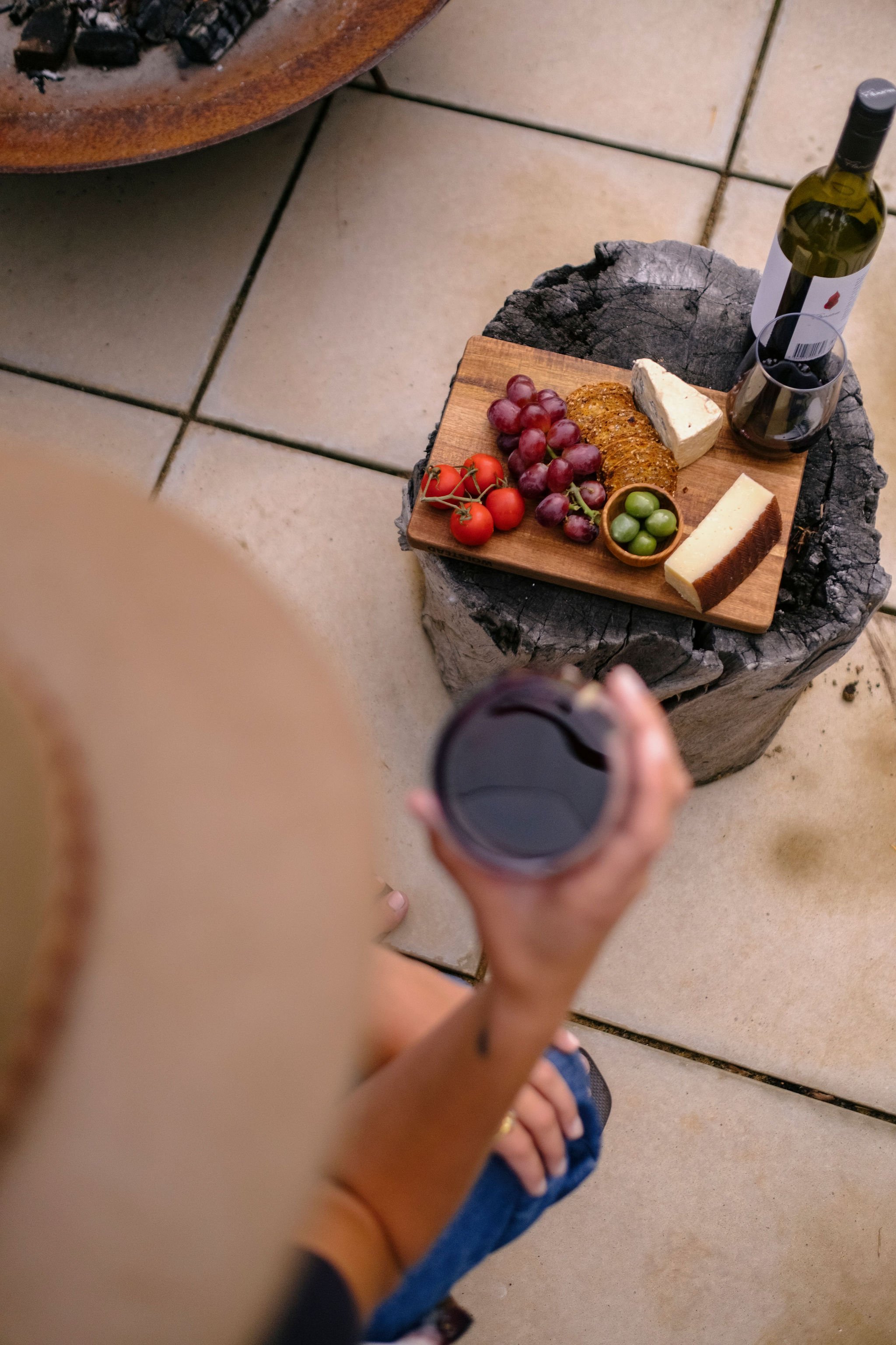 A person holding a glass of red wine, with a cheese and fruit platter on a wooden slab in front of them on a tiled floor.
