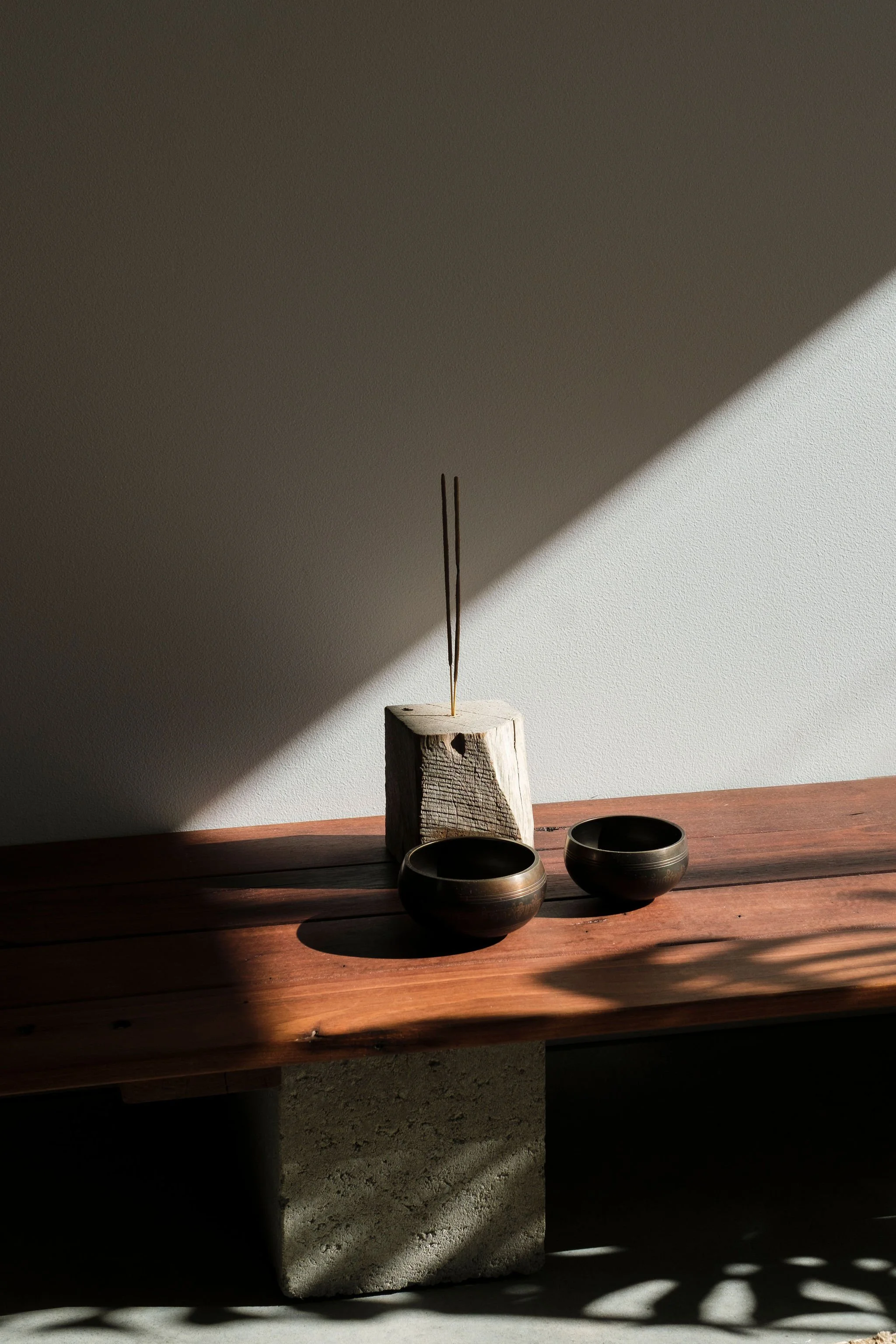 A minimalist tabletop arrangement featuring two black bowls, a wooden block, and two incense sticks casting shadows on a surface with light and shadow contrast.