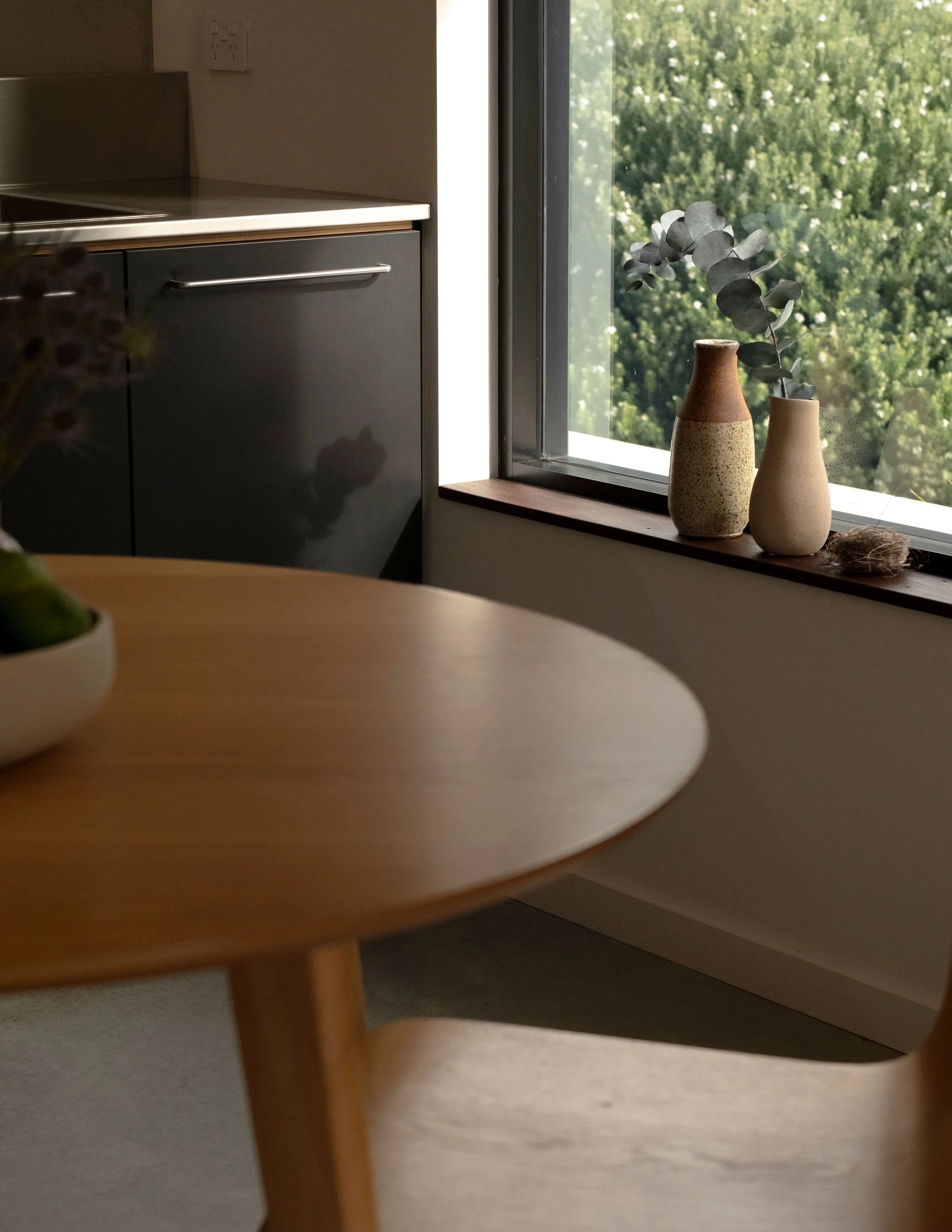 A cozy room corner with a large window overlooking green trees, decorated with two ceramic vases containing eucalyptus leaves, and a small bird's nest.