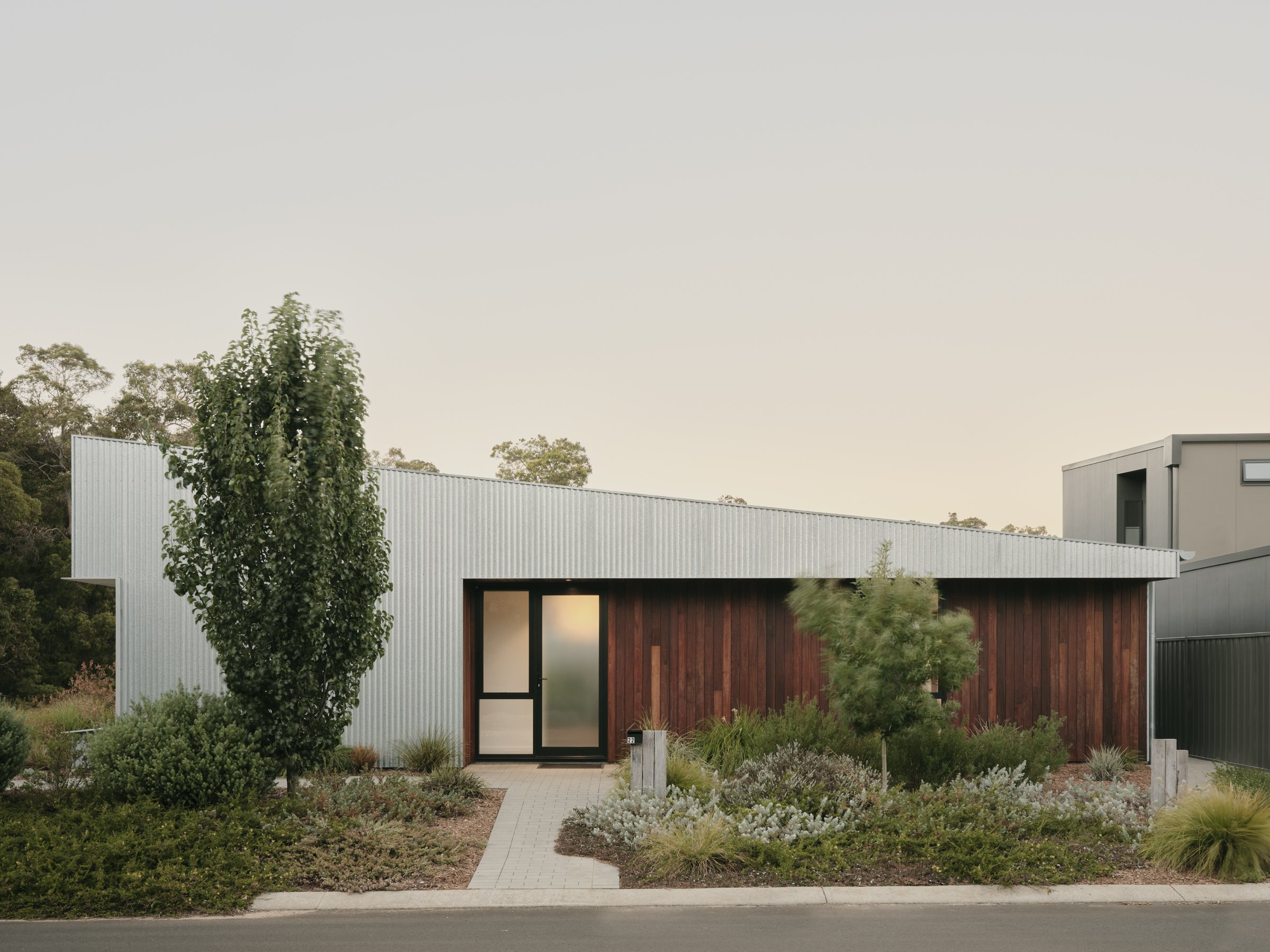 Modern house with galvanised metal and timber cladding and large windows, surrounded by landscaped garden with grasses and shrubs.
