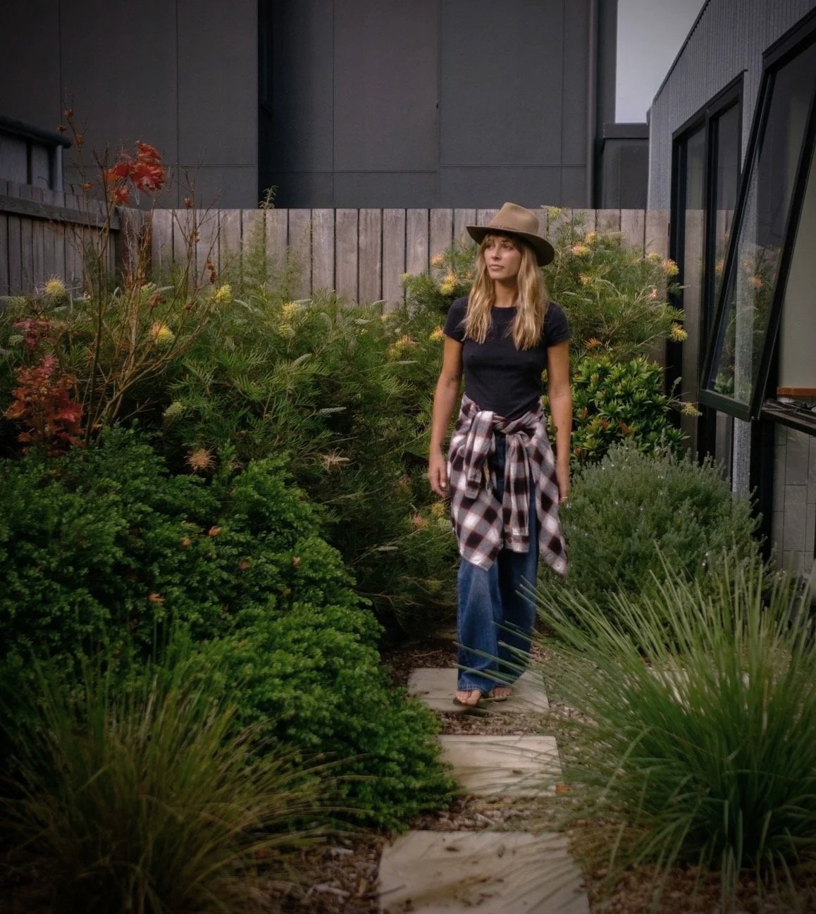 A woman wearing a wide-brimmed hat, black t-shirt, plaid shirt tied around her waist, and wide-leg jeans standing on stepping stones in a lush garden.