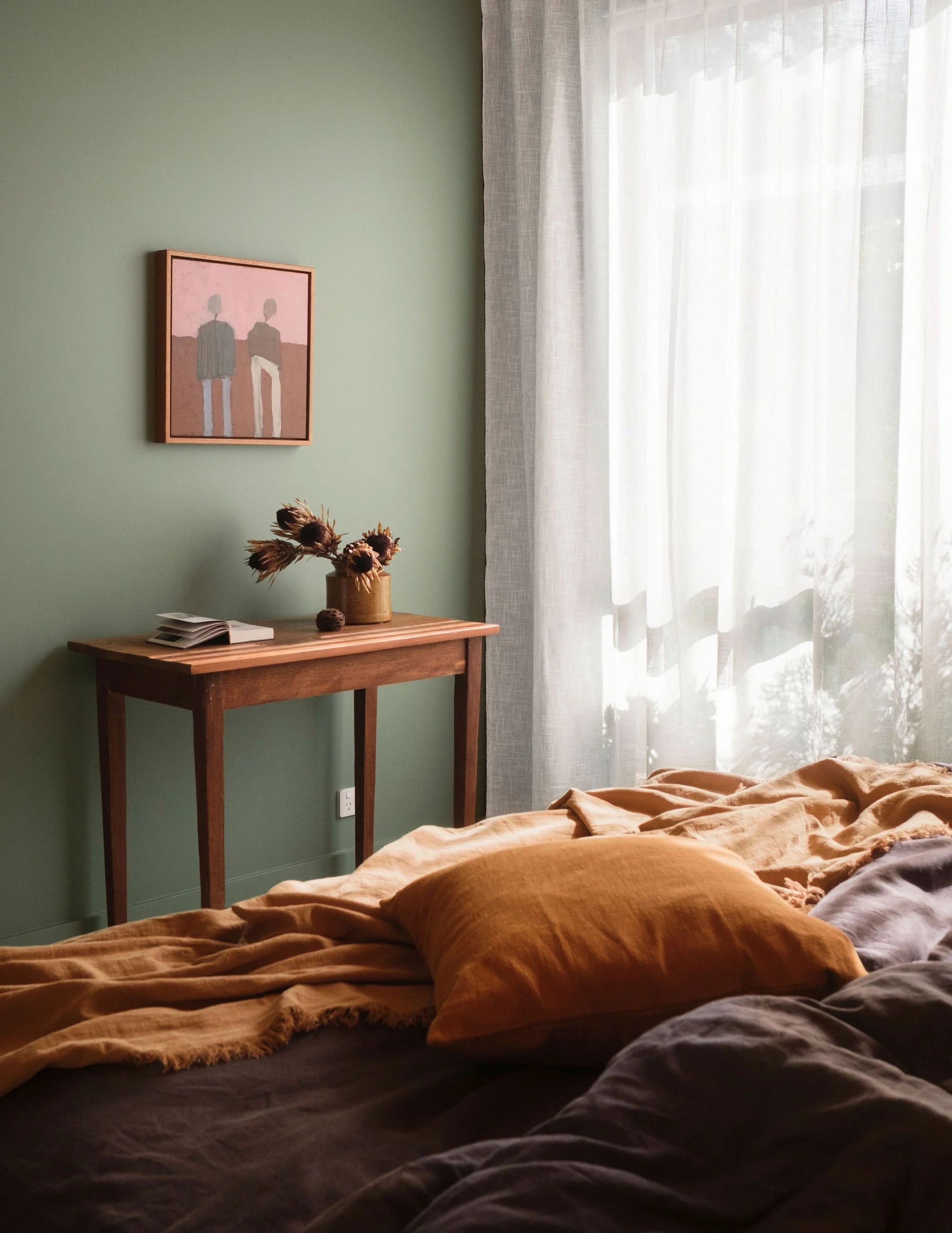 A cozy bedroom with a green wall, a wooden side table, and a bed with brown and gray blankets. A sunflower arrangement is on the side table, with a framed painting of two abstract figures on the wall. Sheer white curtains are on the window, allowing natural light into the room.