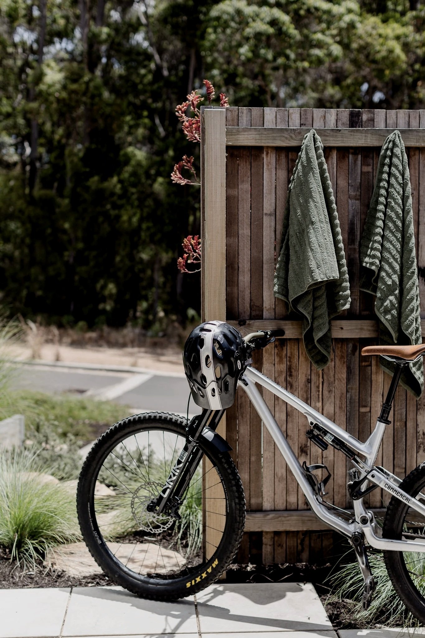 A white mountain bike with a black helmet hanging on the handlebar, leaned against a wooden fence. Two green towels are draped over the fence, with a small plant with pinkish buds visible behind it. The background shows trees and bushes, indicating an outdoor setting.