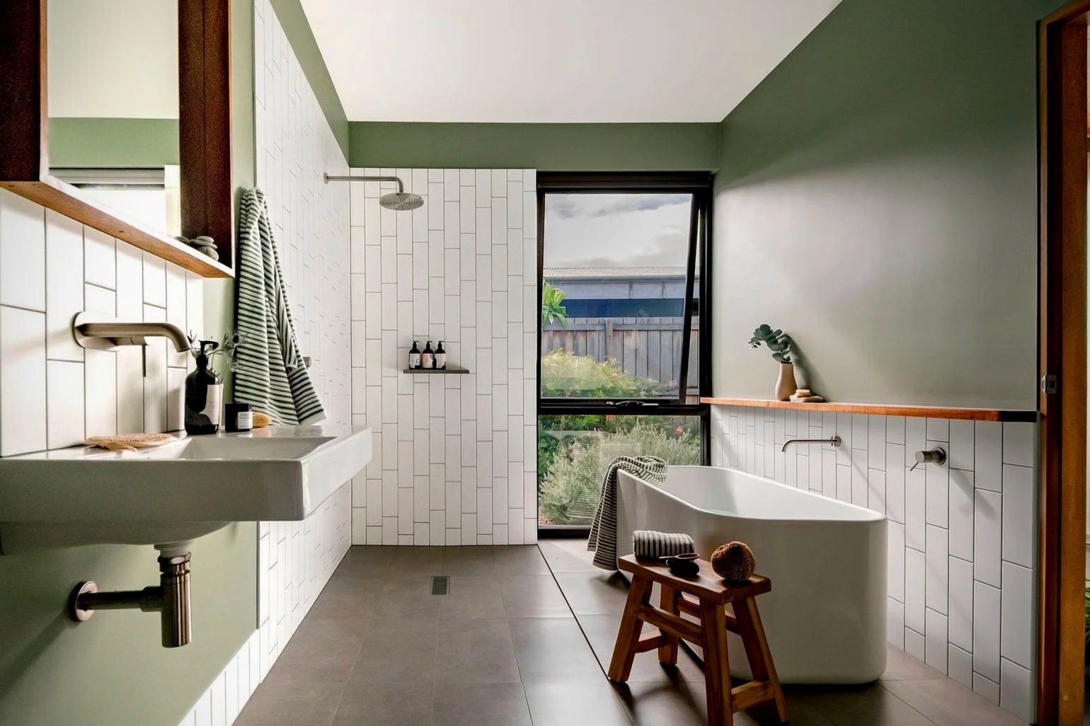 Modern bathroom with green and white walls, a large window overlooking a garden, a white bathtub with a wooden stool and a towel, a white sink with a striped towel, a showerhead, and decorative plants.