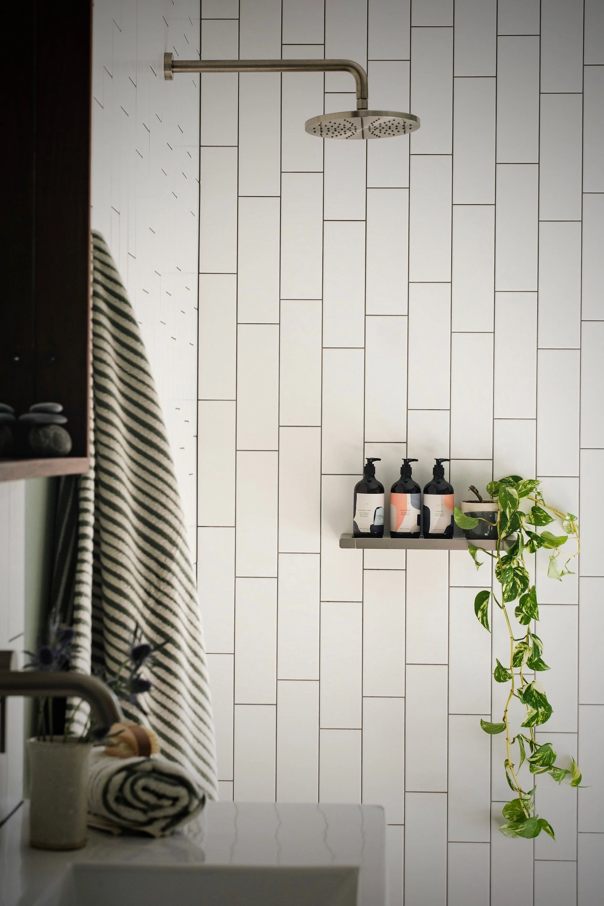 Bathroom with white subway tile walls, a round showerhead on a metal pipe, three black bottles with pump dispensers on a gray shelf, a small potted plant, and a striped towel hanging on a hook. The photo is taken in a mirror, reflecting the scene.