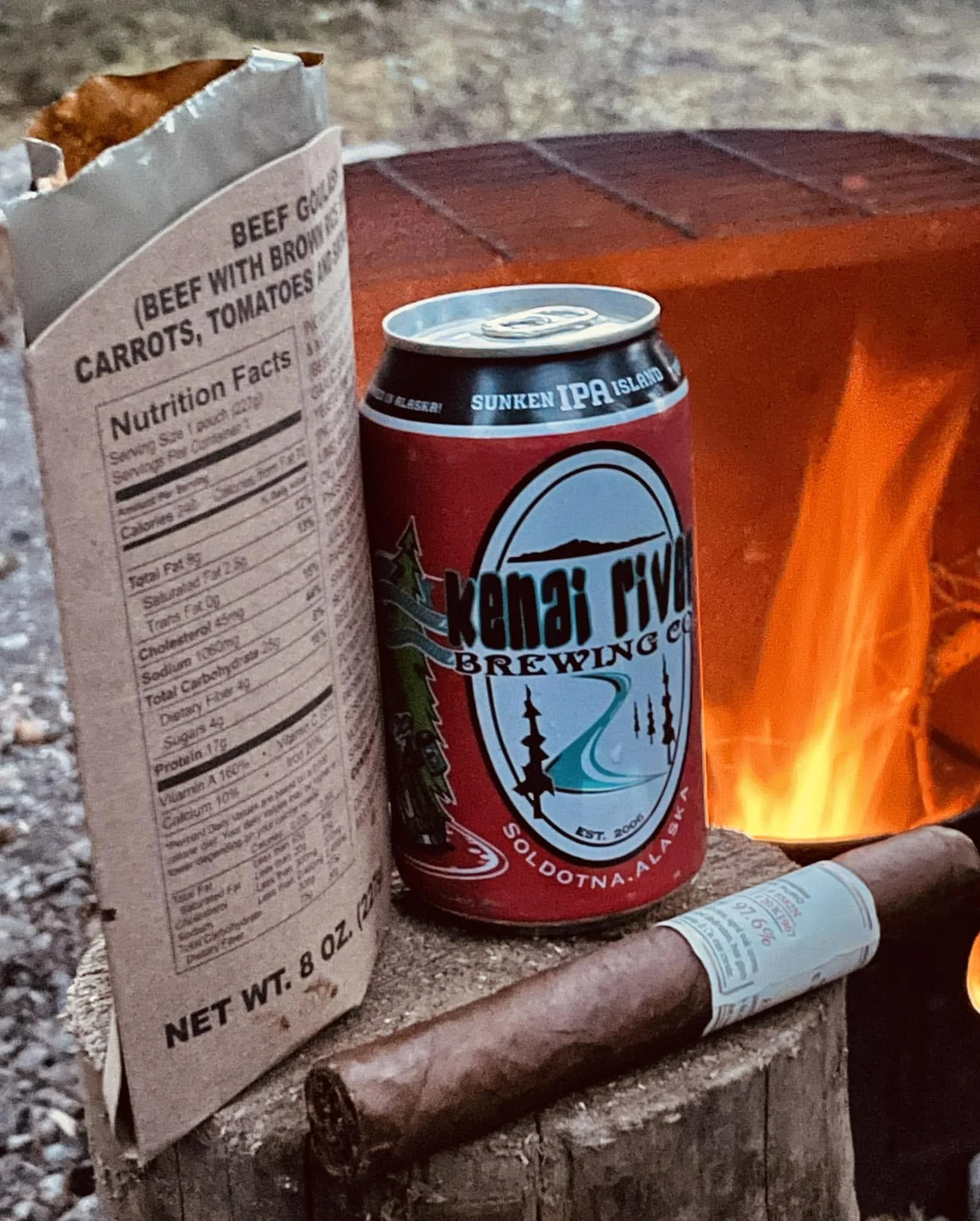 A cigar beside a campfire with a field meal and Alaska-brewed beer.