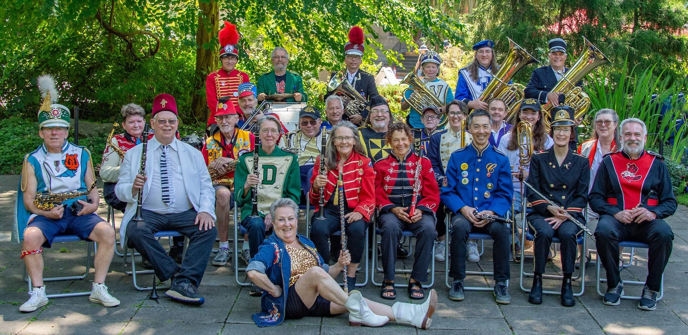 Group of people dressed in colorful marching band uniforms, some holding musical instruments, posing outdoors under trees.