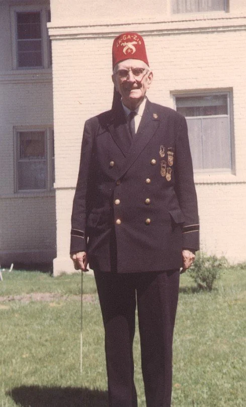 An elderly man in a formal military uniform with medals, wearing a red fez hat, standing outside in front of a building with grass and bushes.