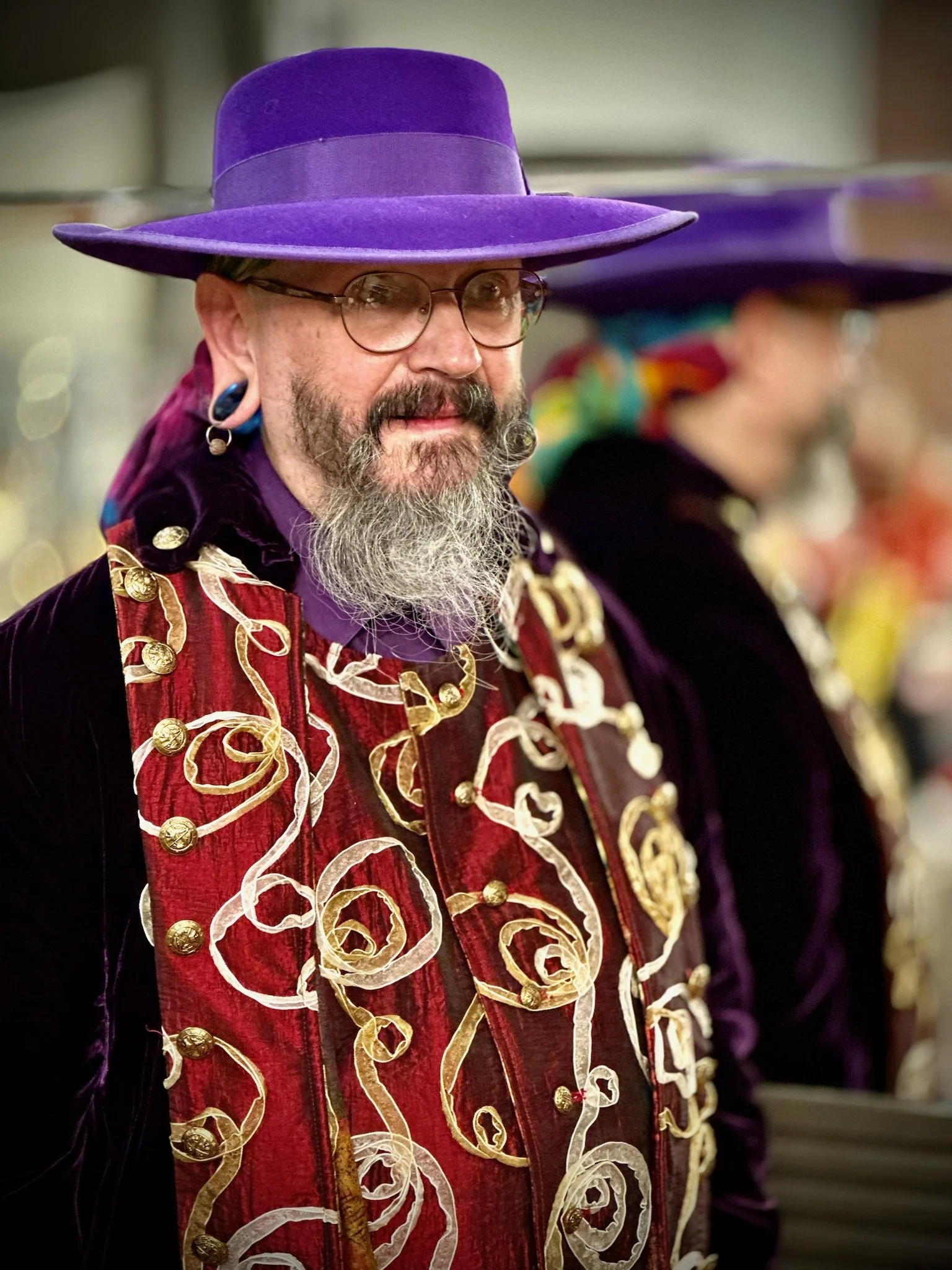 A man wearing a purple hat and glasses, dressed in a red and black elaborate costume with gold accents, standing in a line with other similarly dressed individuals.