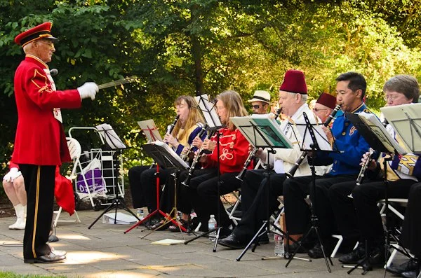 A groups of musicians playing their instruments outdoors, led by a band conductor in a red uniform with a hat. The musicians are sitting in a row, reading sheet music, with trees and sunlight in the background.