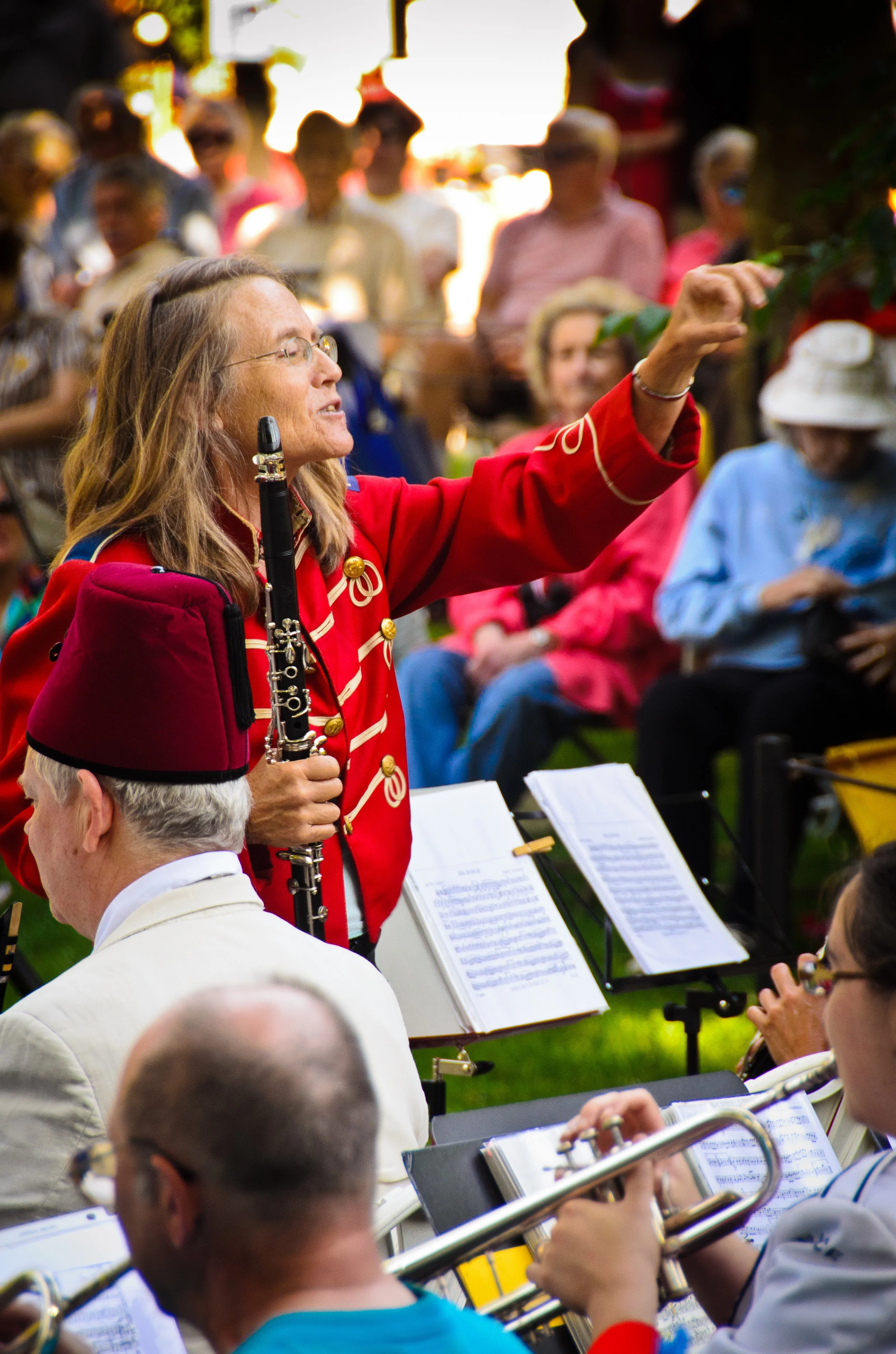 A woman in a red jacket with gold embellishments, holding a clarinet, conducting an outdoor music performance in front of an audience of seated people.