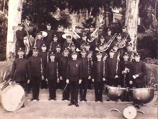 A vintage black-and-white photograph of a music band with young men in dark suits, standing outdoors with trees in the background. They hold brass instruments like trumpets, trombones, and saxophones, with some percussion instruments in front.