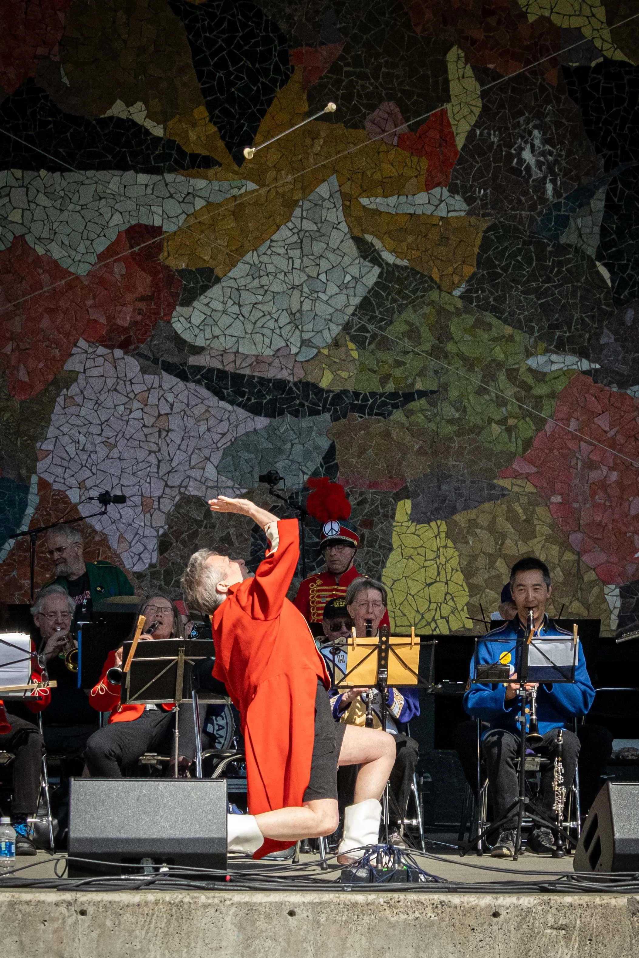 A group of musicians performing on an outdoor stage with a colorful mosaic mural as the backdrop. Some members are playing instruments, and a conductor in an orange jacket and white boots is leading the group.
