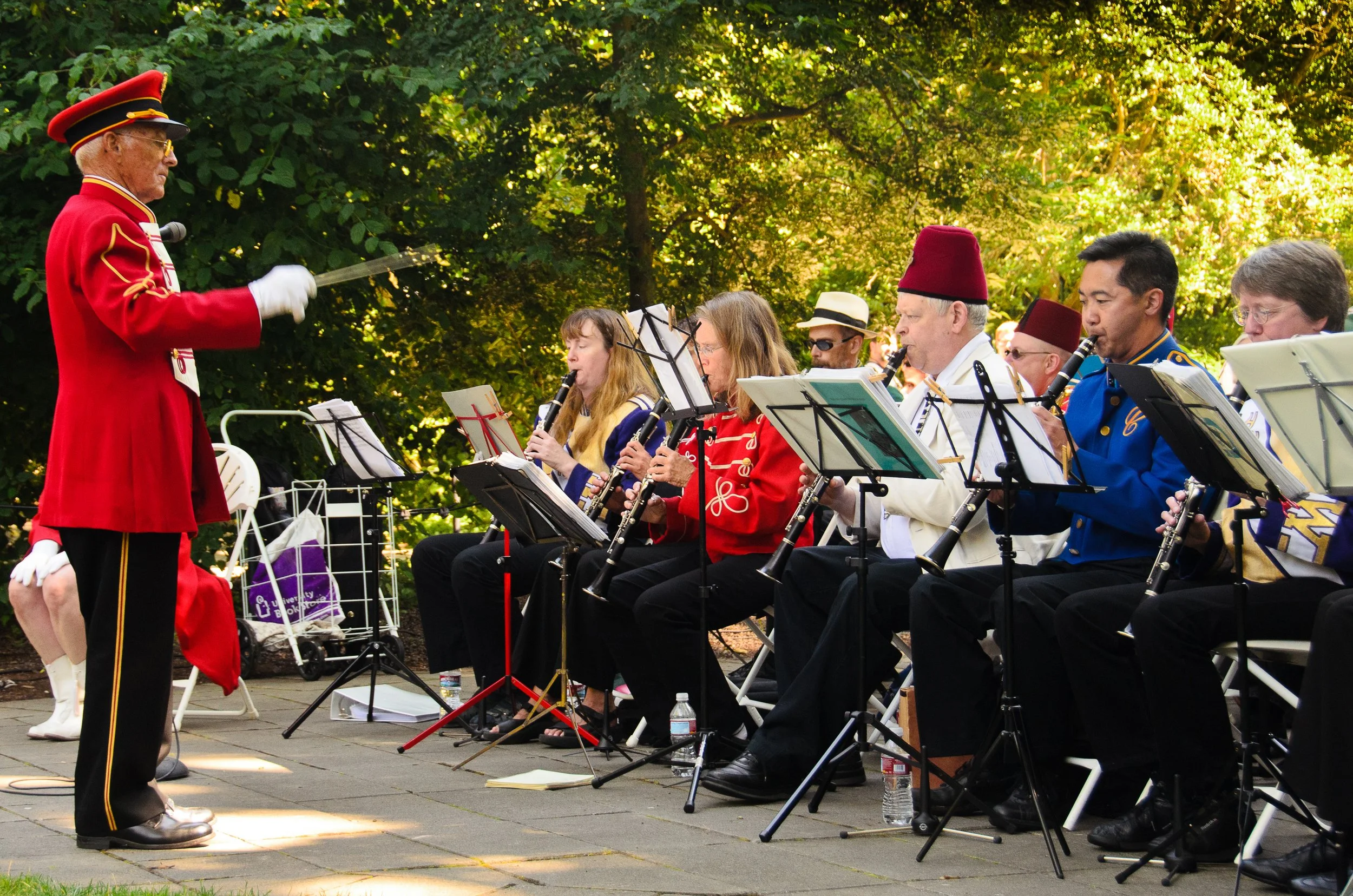 A marching band performing outdoors with a conductor leading. The band members are playing wind instruments and dressed in colorful uniforms, some wearing hats. There is greenery in the background and sunlight filtering through the trees.