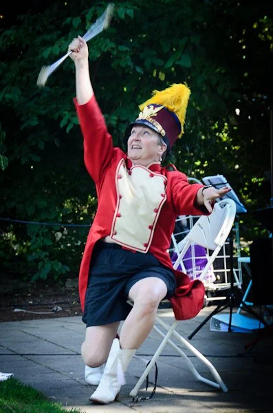 A woman dressed as a marching band member, wearing a red coat, beige vest, and a yellow feathered hat, sits on a folding chair outdoors amid green trees, holding a baton and a smartphone, with an excited expression.