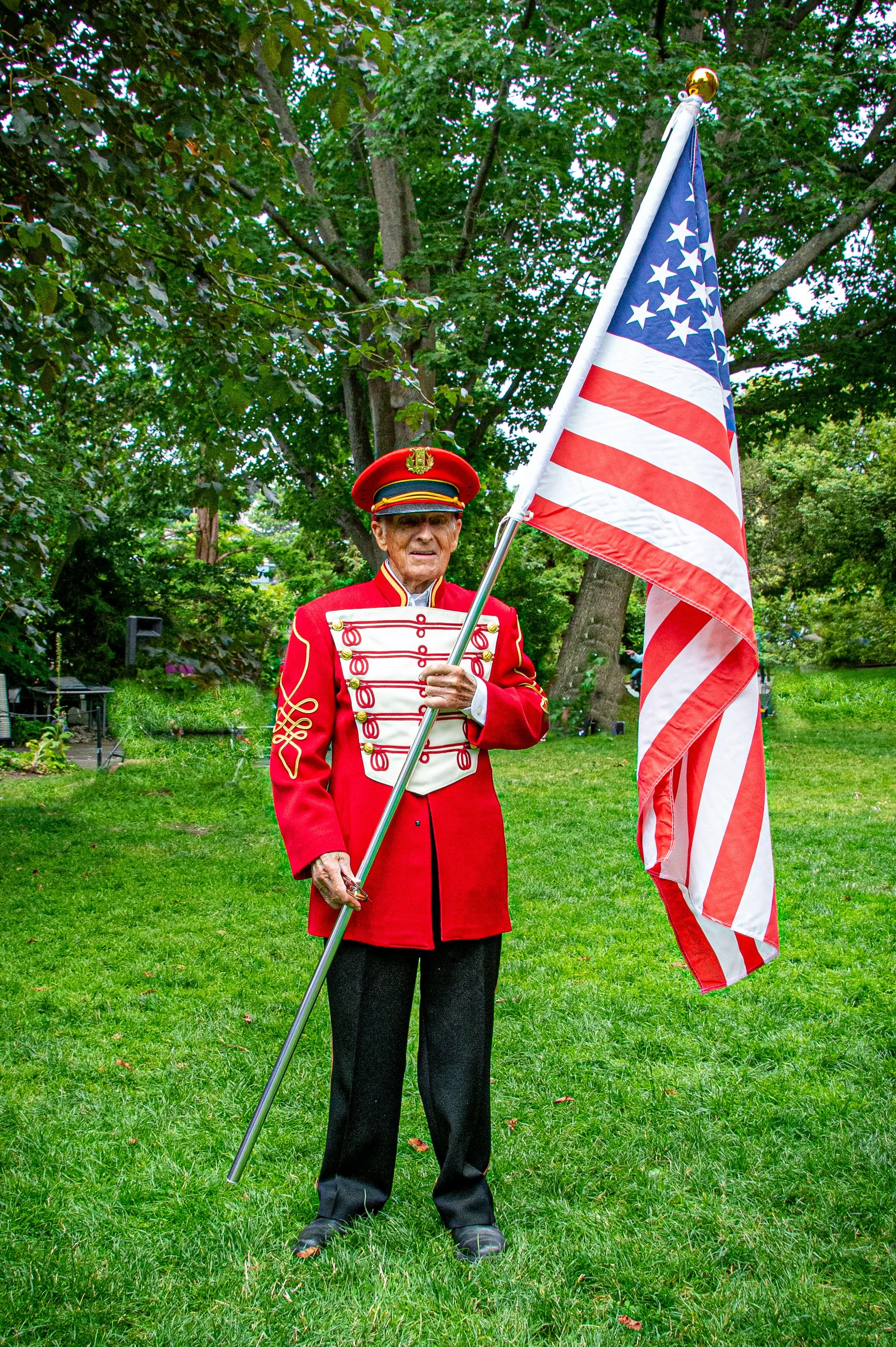 An elderly man dressed in a red, white, and gold American Revolutionary War uniform holding an American flag outdoors with trees in the background.