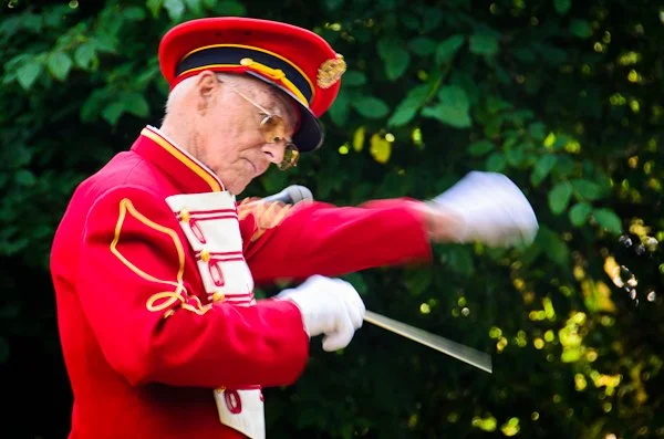 A man dressed in a red military band uniform with gold accents, a red cap with black and gold trim, performing a conductorial gesture with a baton in front of green leafy foliage.