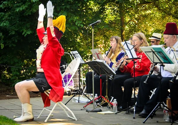 A person in a red military-style jacket, white gloves, and a yellow feathered hat performing as a drummer in front of a seated band playing wind instruments outdoors.