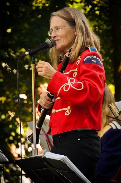 A woman in a red band jacket with a patch reading 'AUBURN' and a military insignia, singing into a microphone outdoors during the daytime.
