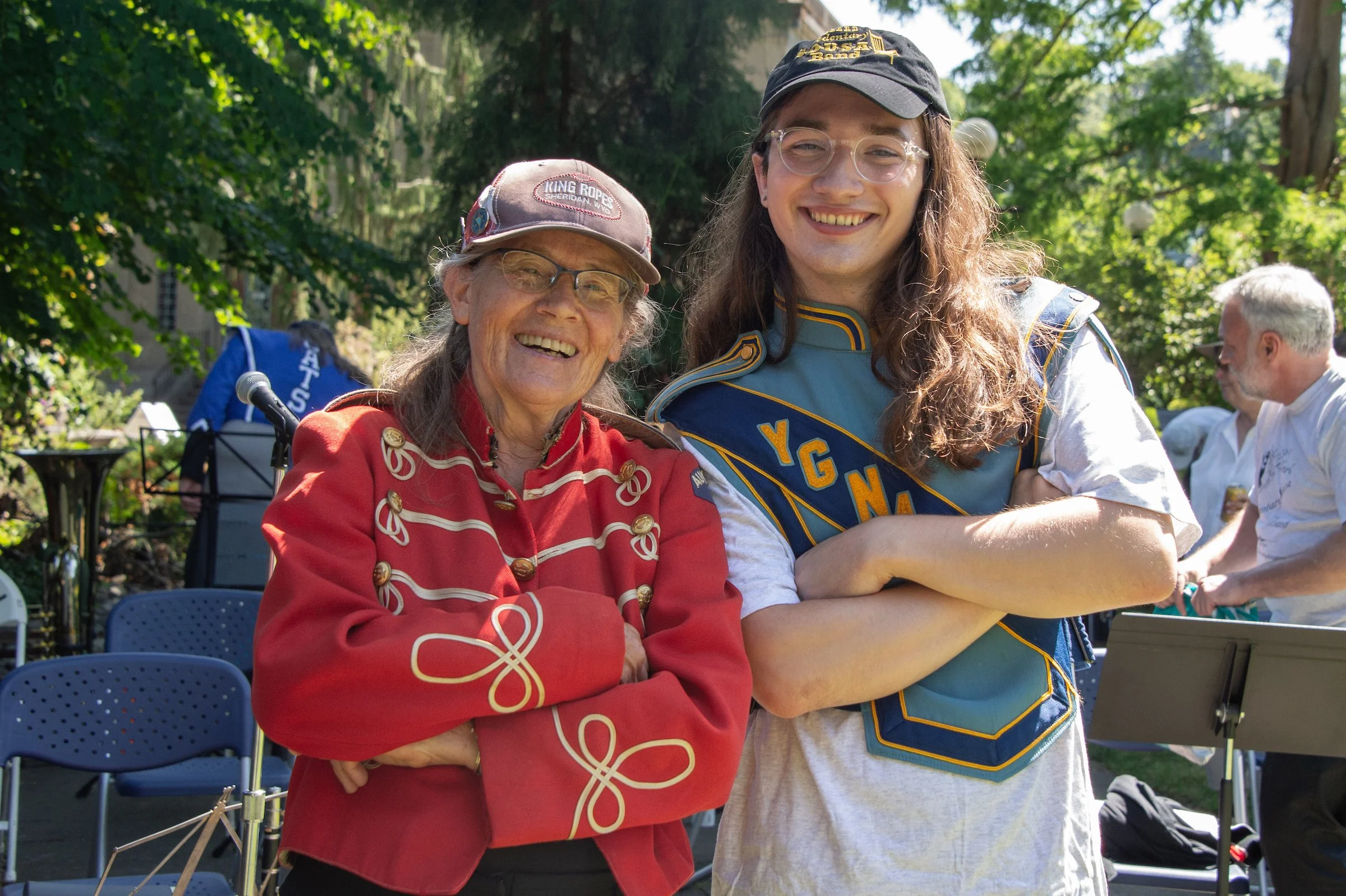 Two women smiling in an outdoor park, one wearing a red jacket with decorative gold buttons and a cap, the other in a blue sash and a black cap, with green trees and other people in the background.