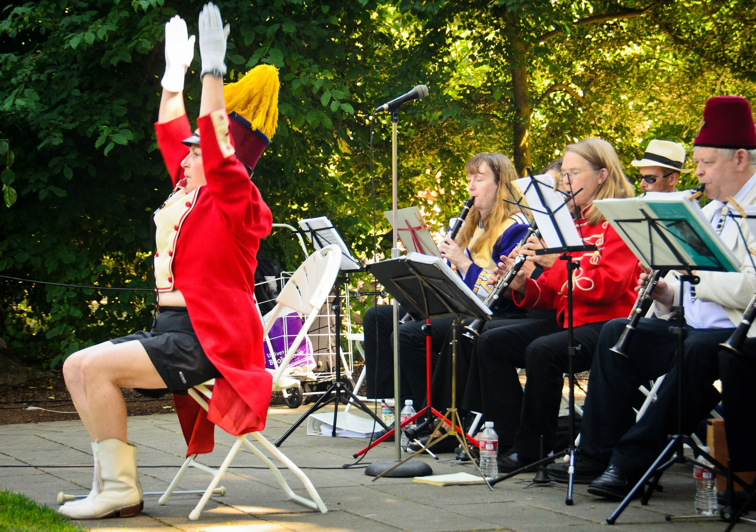 A band performing outdoors, with musicians playing wind instruments and a conductor in a red jacket and a yellow feathered hat leading them. The scene is set in a park with trees in the background.