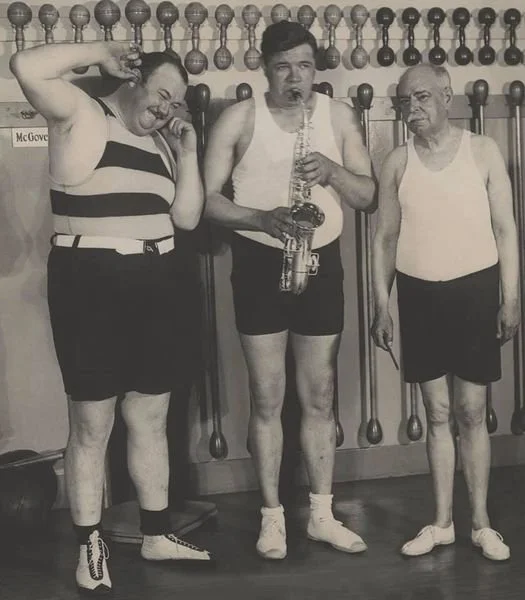 Three men standing in front of a wall with many bowling balls, one man is playing a saxophone, and all are dressed in vintage athletic clothing.