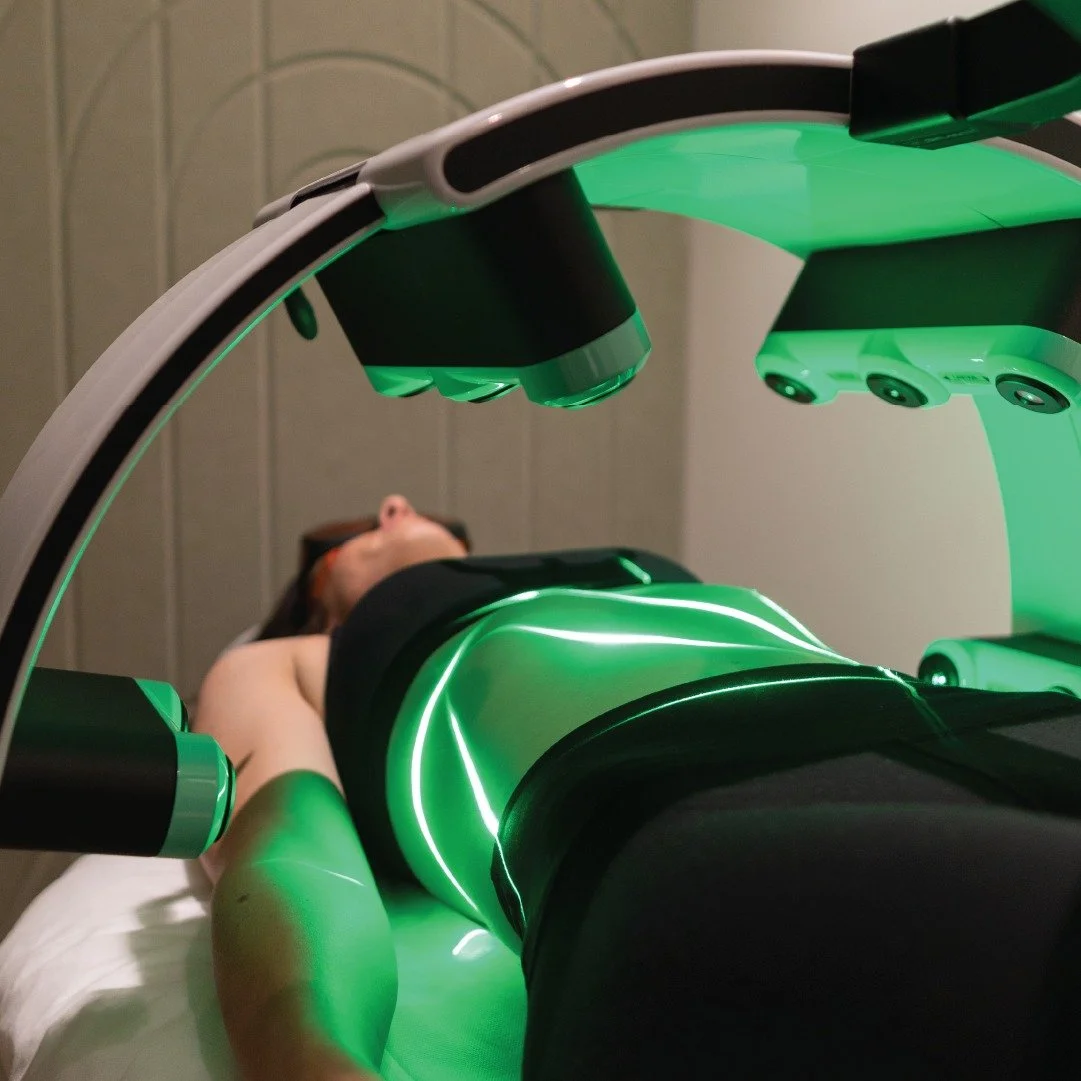 Client lying on a treatment table beneath an Erchonia Emerald Laser device emitting green low-level laser light in a clinical setting.