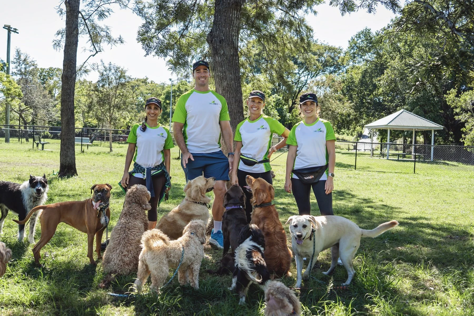 Four people, two women and two men, standing outdoors in a park with many dogs. The people are wearing white and green shirts with a logo that says 'SAS Dogs.' The dogs are a variety of breeds and sizes, some sitting and some standing on the grass. The background includes trees, a fenced area, a small pavilion, and picnic tables, with bright sunlight and clear skies.