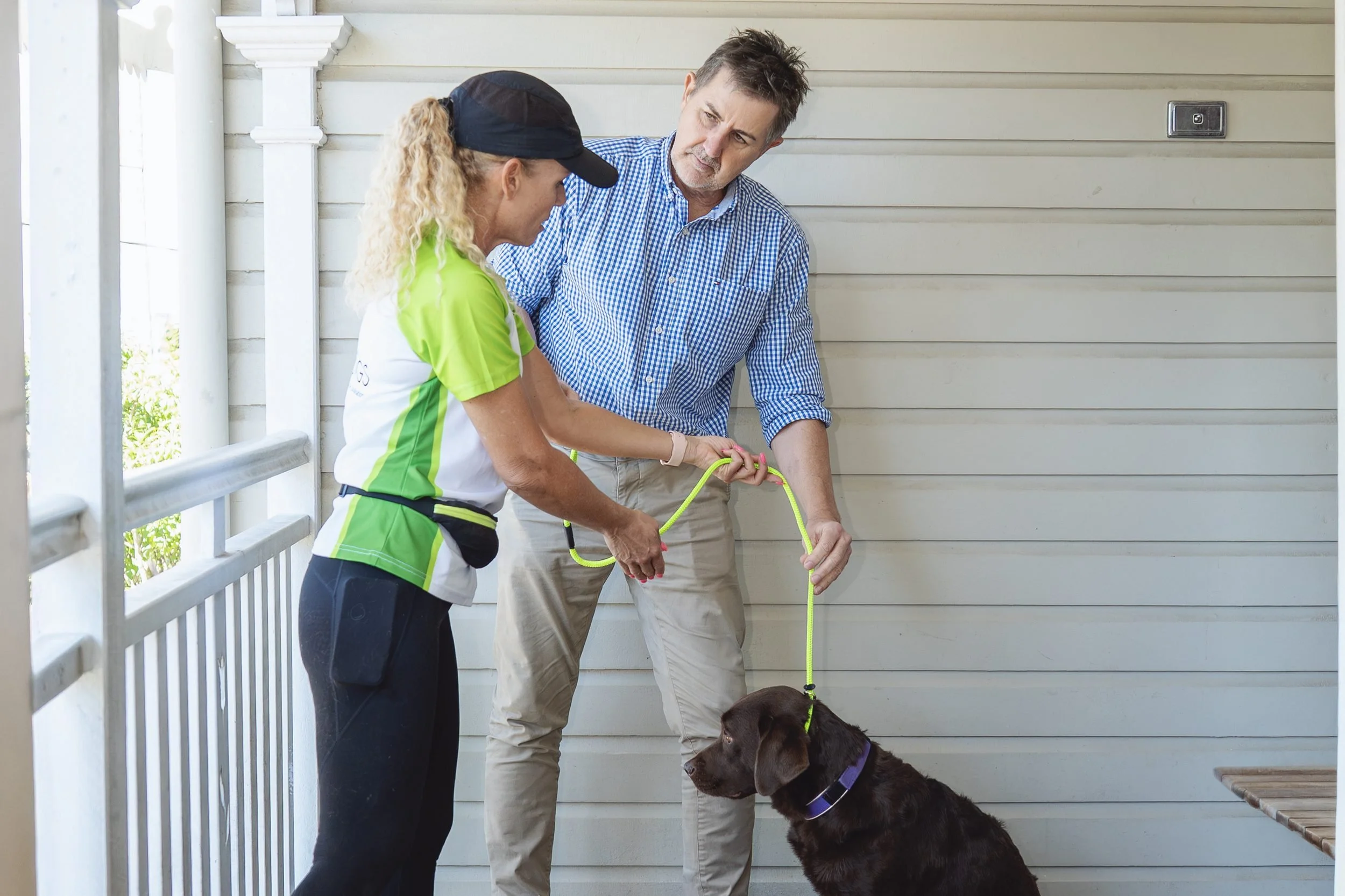 A woman and a man with a brown dog are standing on a porch, practicing a training command with the dog. The woman is wearing a black hat, bright green athletic shirt, and black pants, while the man is dressed in a blue checkered shirt and beige pants. The woman is holding a yellow leash and appears to be giving commands, while the man watches attentively.