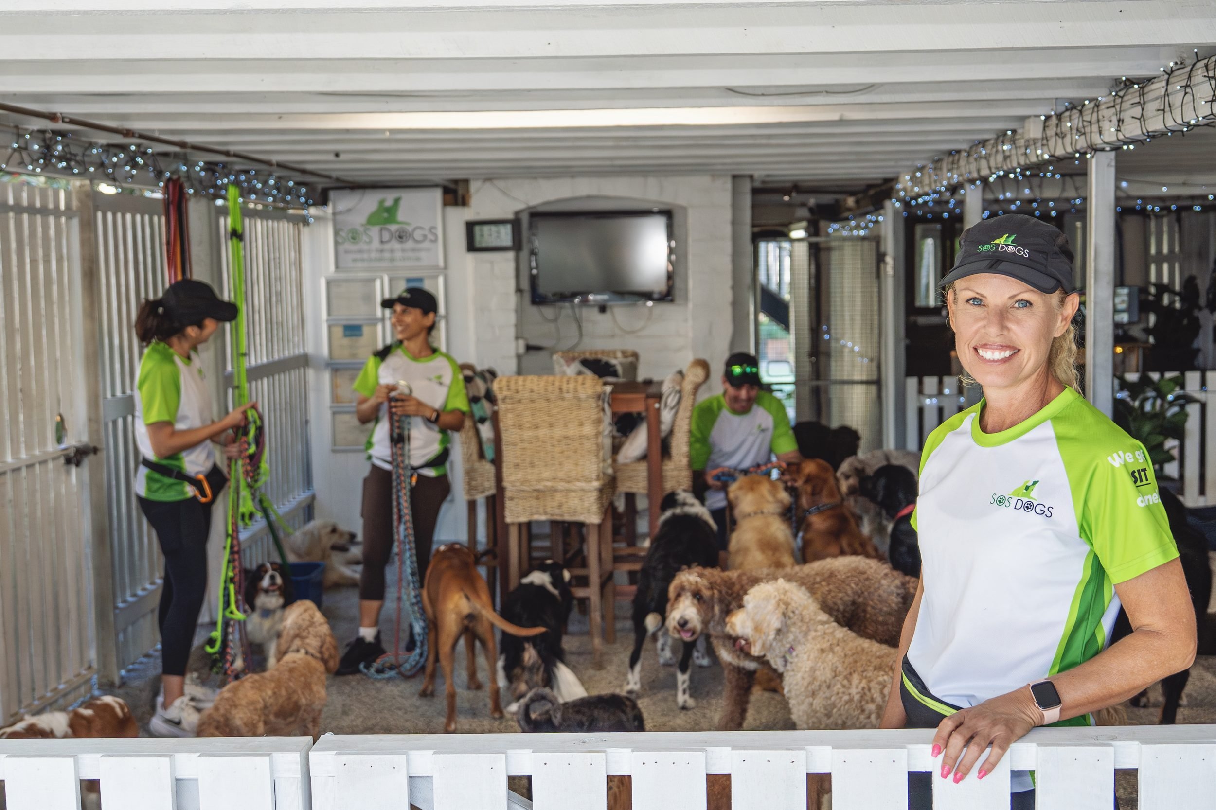 A group of women and a man in bright green and white shirts with a logo that says 'SoS Dogs' are interacting with a large group of dogs in an indoor setting. The woman in the foreground is smiling and wearing a black cap with the same logo. The background features string lights, a television, and various shelter furniture.