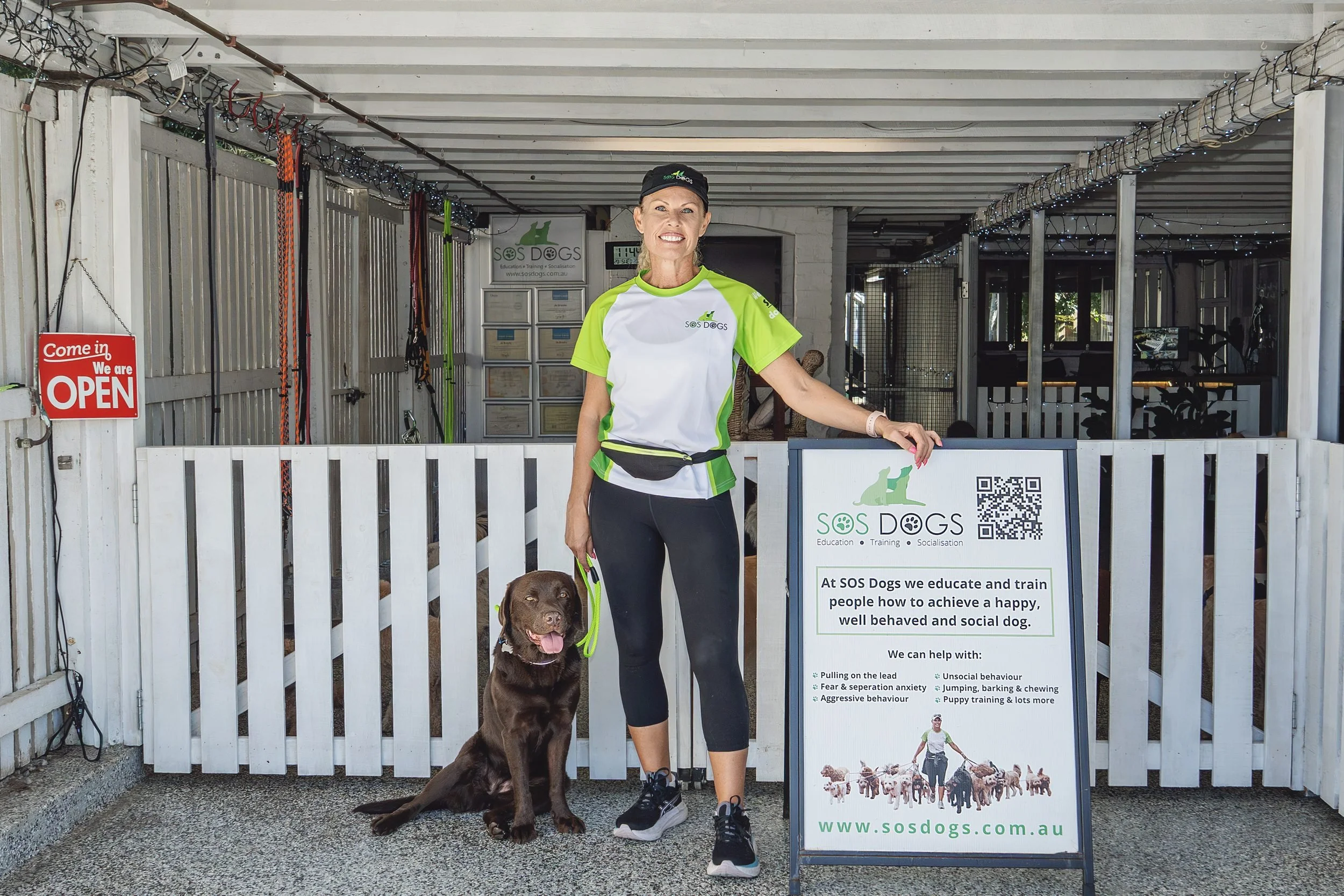 A woman in a green and white SOS Dogs training shirt and black cap standing with a chocolate Labrador Retriever dog in front of a white fence and a sign for SOS Dogs, an organization that educates and trains people to manage dog behavior. The woman is smiling and the dog is sitting obediently with its tongue out.