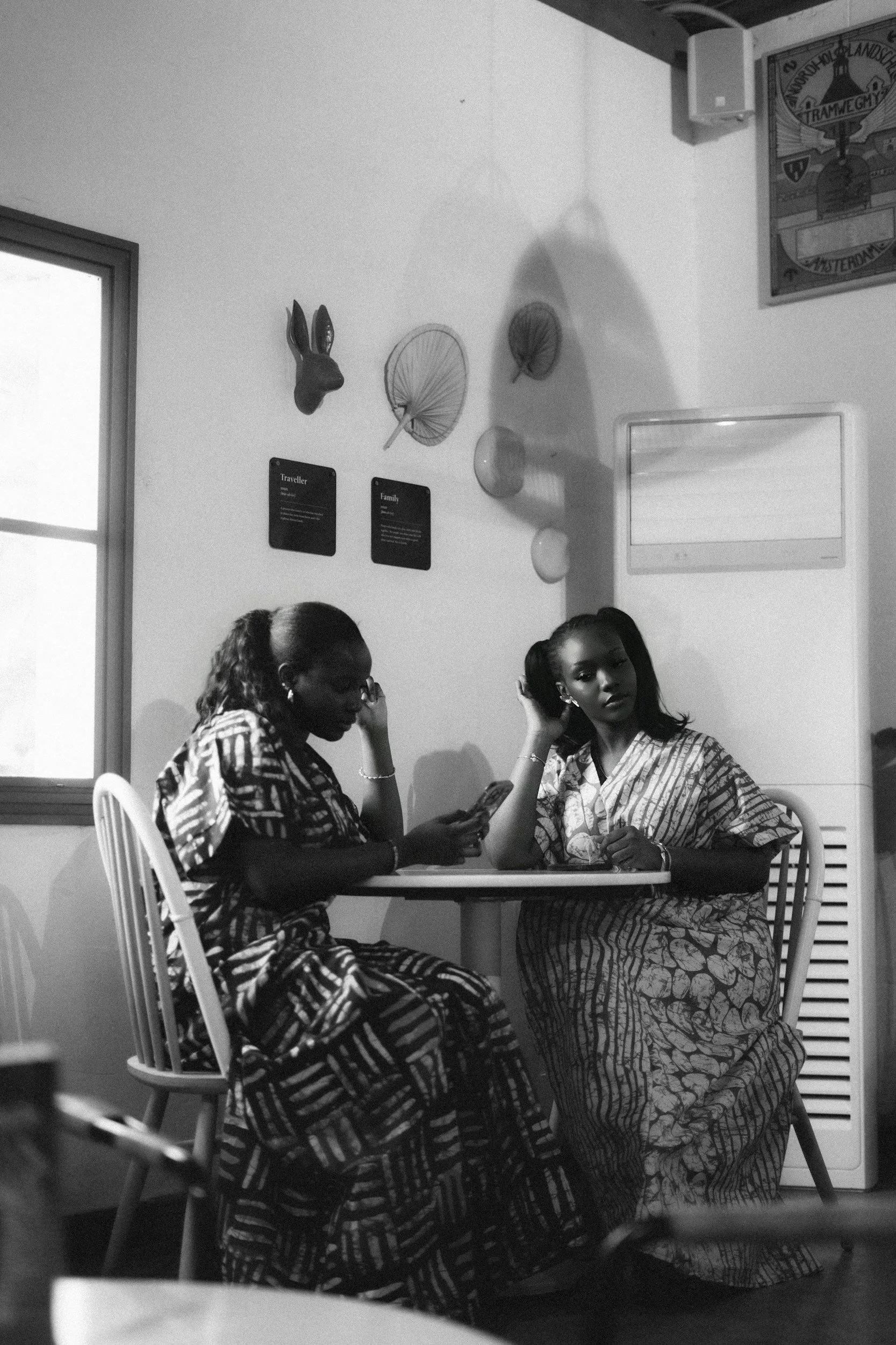 Two women sitting at a small table in a room decorated with wall art, including a rabbit head, umbrellas, and a framed card. One woman is looking at her phone, while the other gazes directly at the camera. Both are dressed in patterned clothing and have their hair styled in updos.