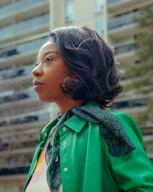 Profile of a woman with short wavy hair, wearing a green jacket and a patterned scarf, standing outdoors with a building in the background.