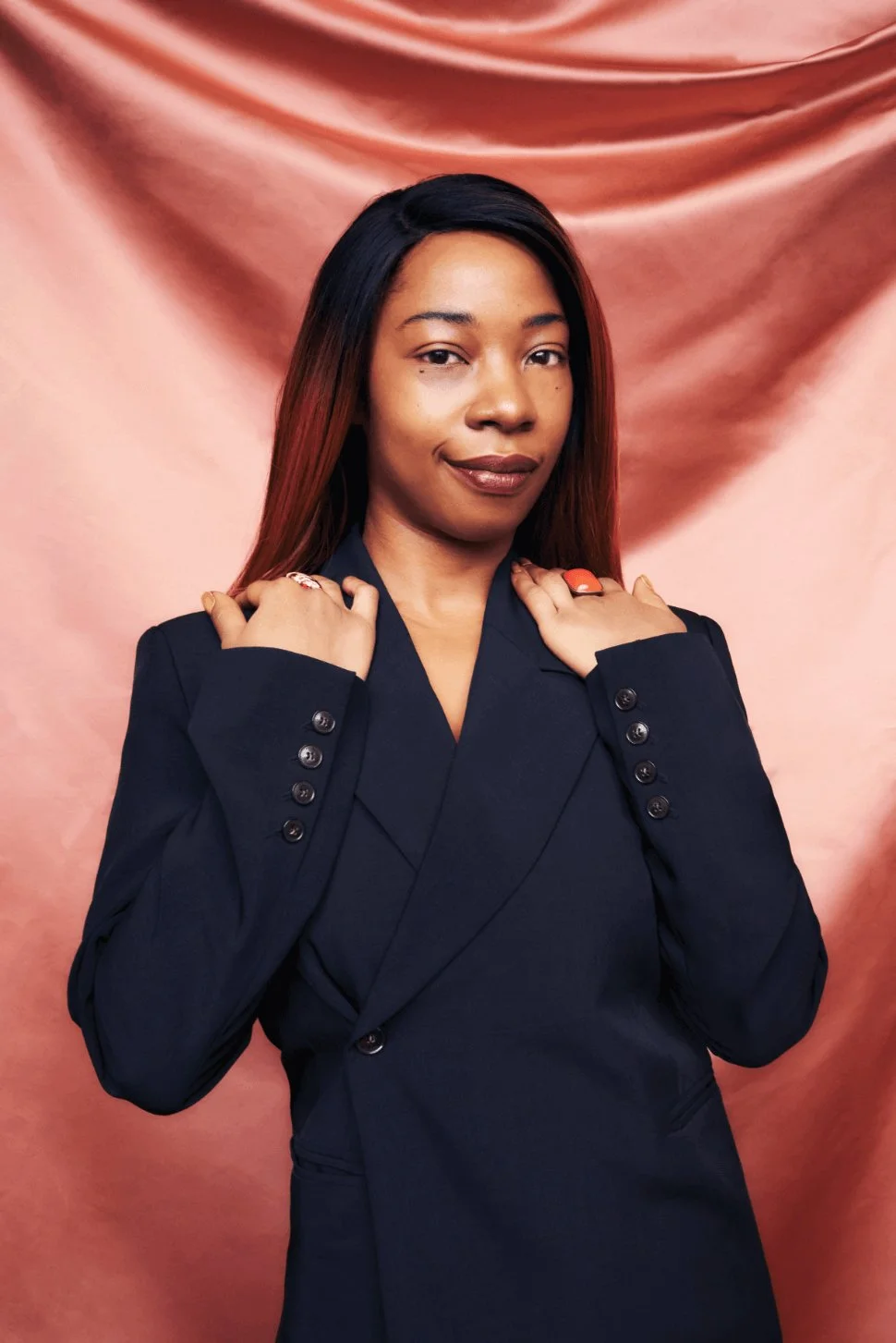 A woman posing against a pink satin backdrop, wearing a black blazer with her hands on her shoulders and a confident expression.