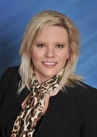 Professional woman with blonde hair wearing a black blazer and a patterned scarf against a blue background.