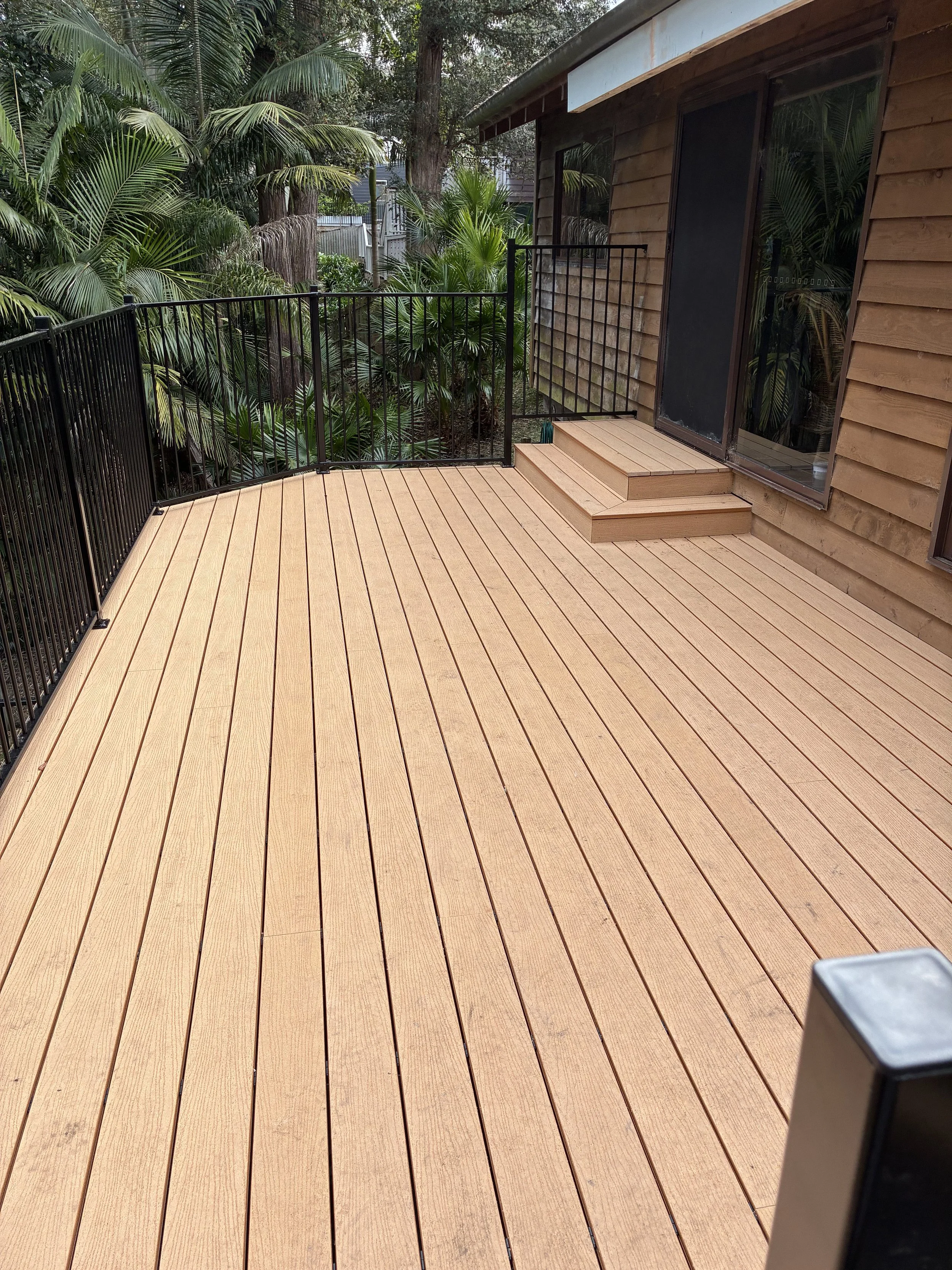 Wooden deck with a black metal railing, sliding glass door, and steps leading to a garden area with various green tropical plants and trees.