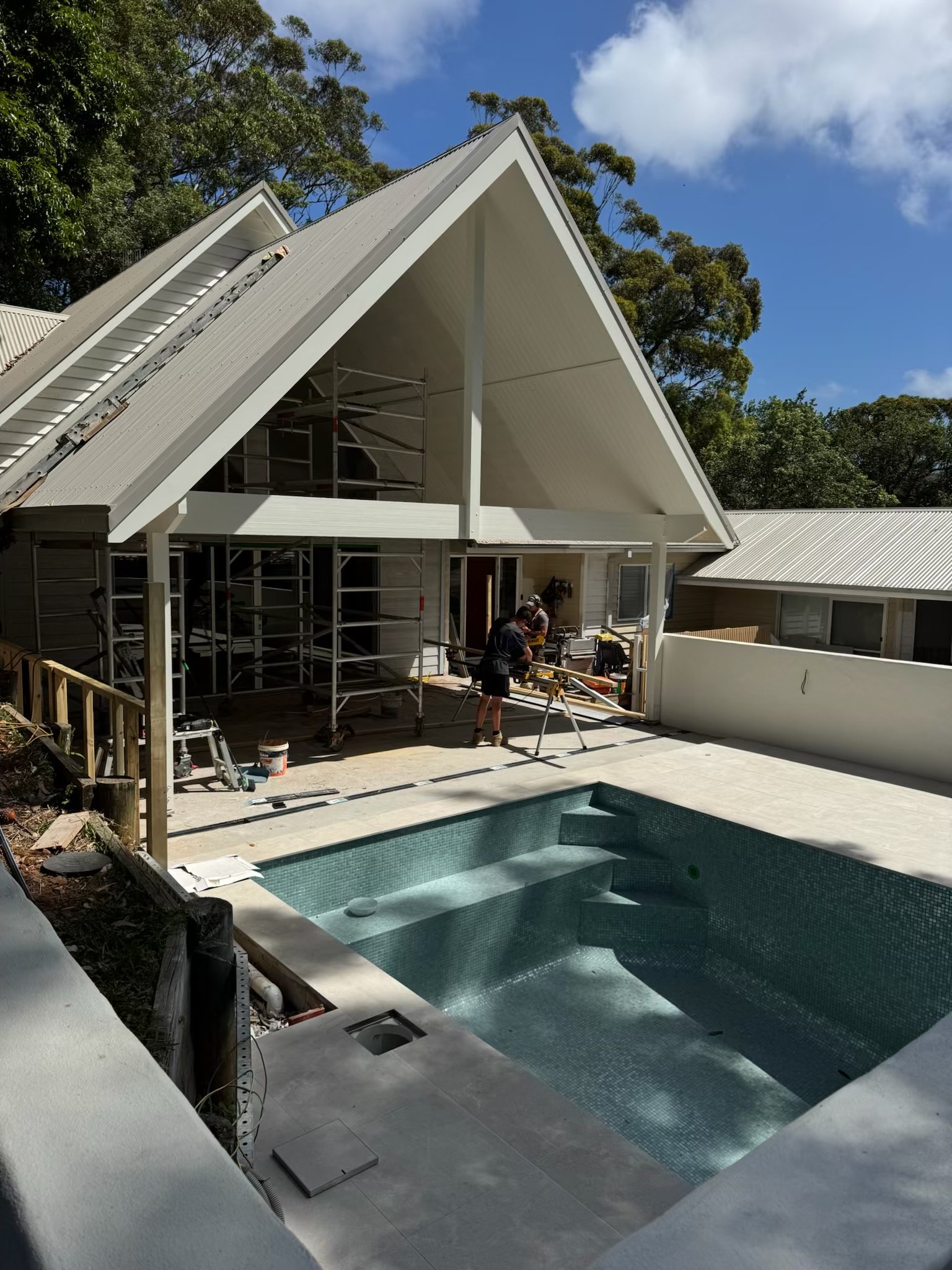 Construction workers working near a swimming pool in a backyard, with a house under construction in the background, scaffolding, and a partly cloudy sky.