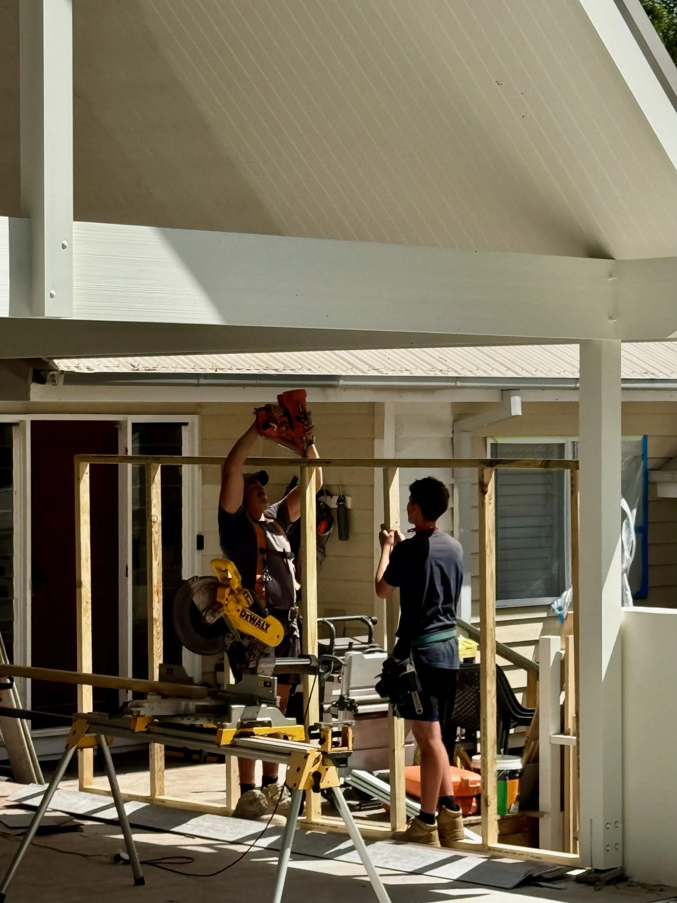 Three builders working on framing a structure outside a house during construction. One man is holding a power tool, while another is standing nearby. A table with tools and materials is in the foreground.