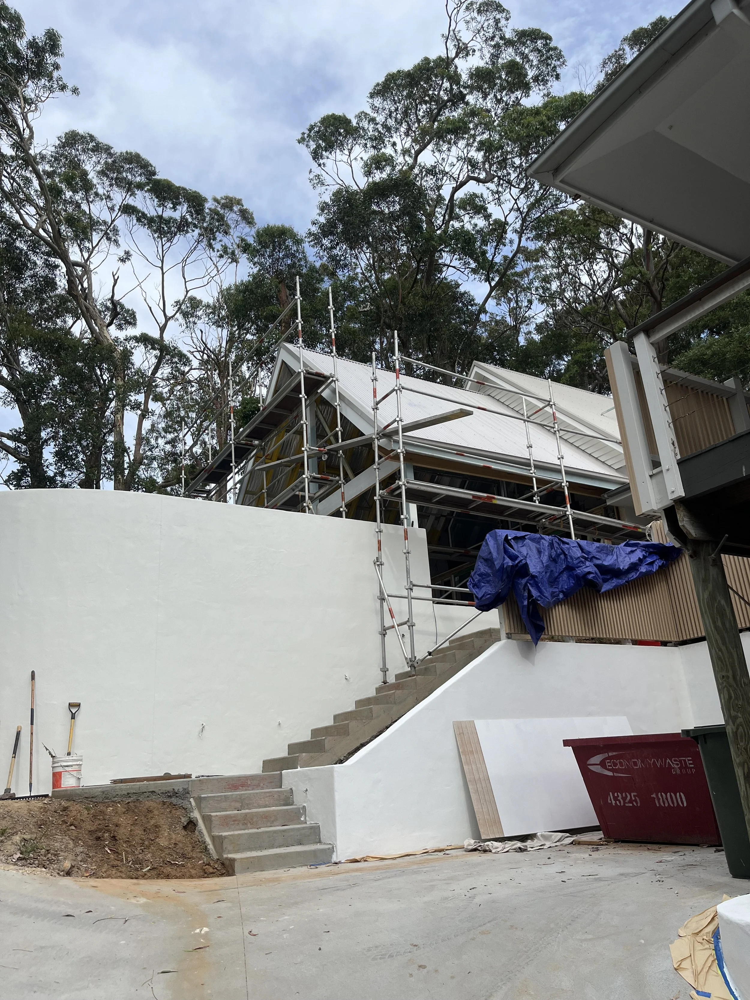 Construction site with a staircase, scaffolding, a partially finished retaining wall, and some construction tools and materials, set outdoors surrounded by tall trees under a partly cloudy sky.