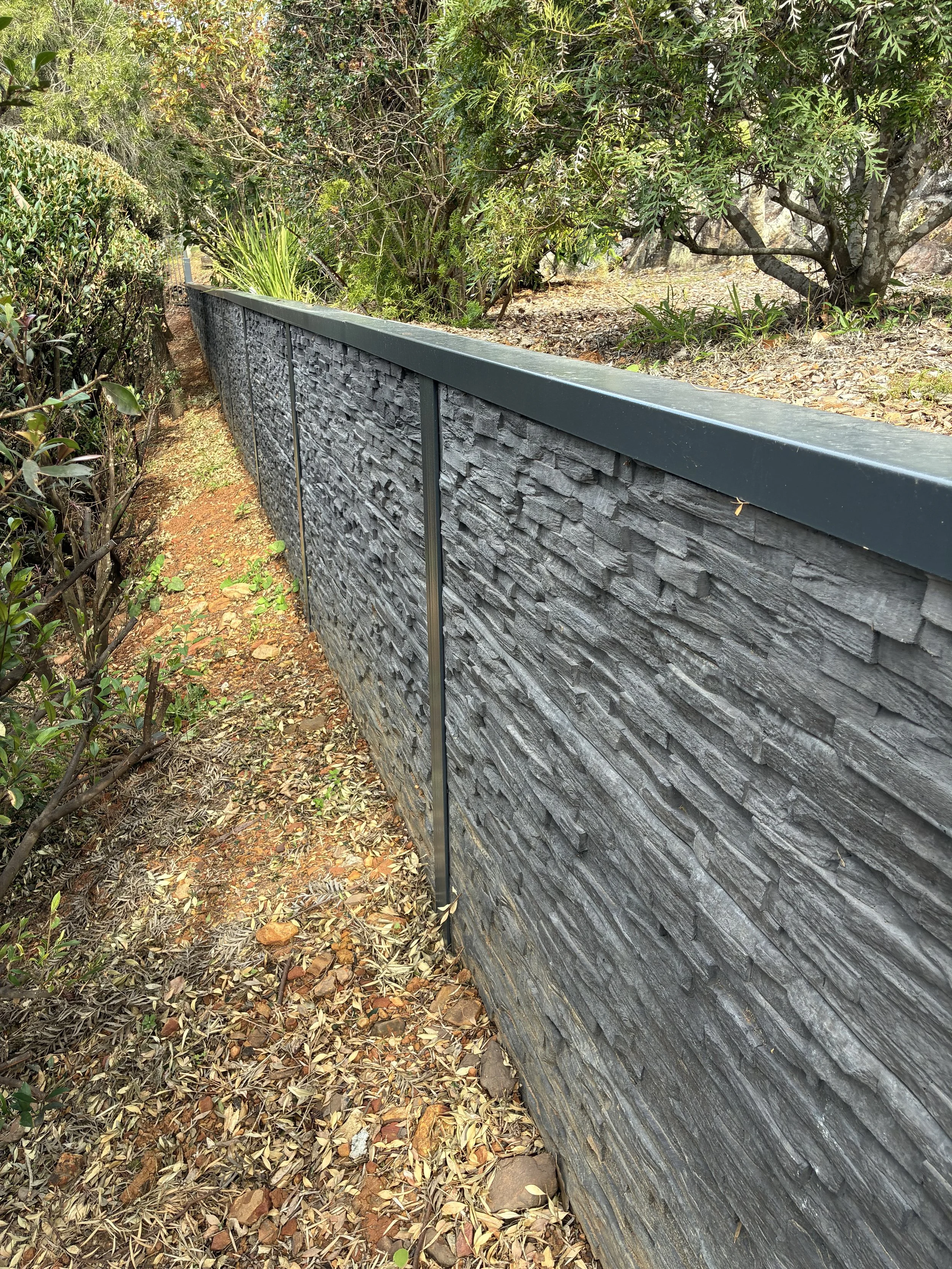 A stone retaining wall with a metal top, situated alongside a narrow dirt walking path. The path is covered with fallen leaves and small rocks. Dense green foliage and trees grow on the other side of the wall.