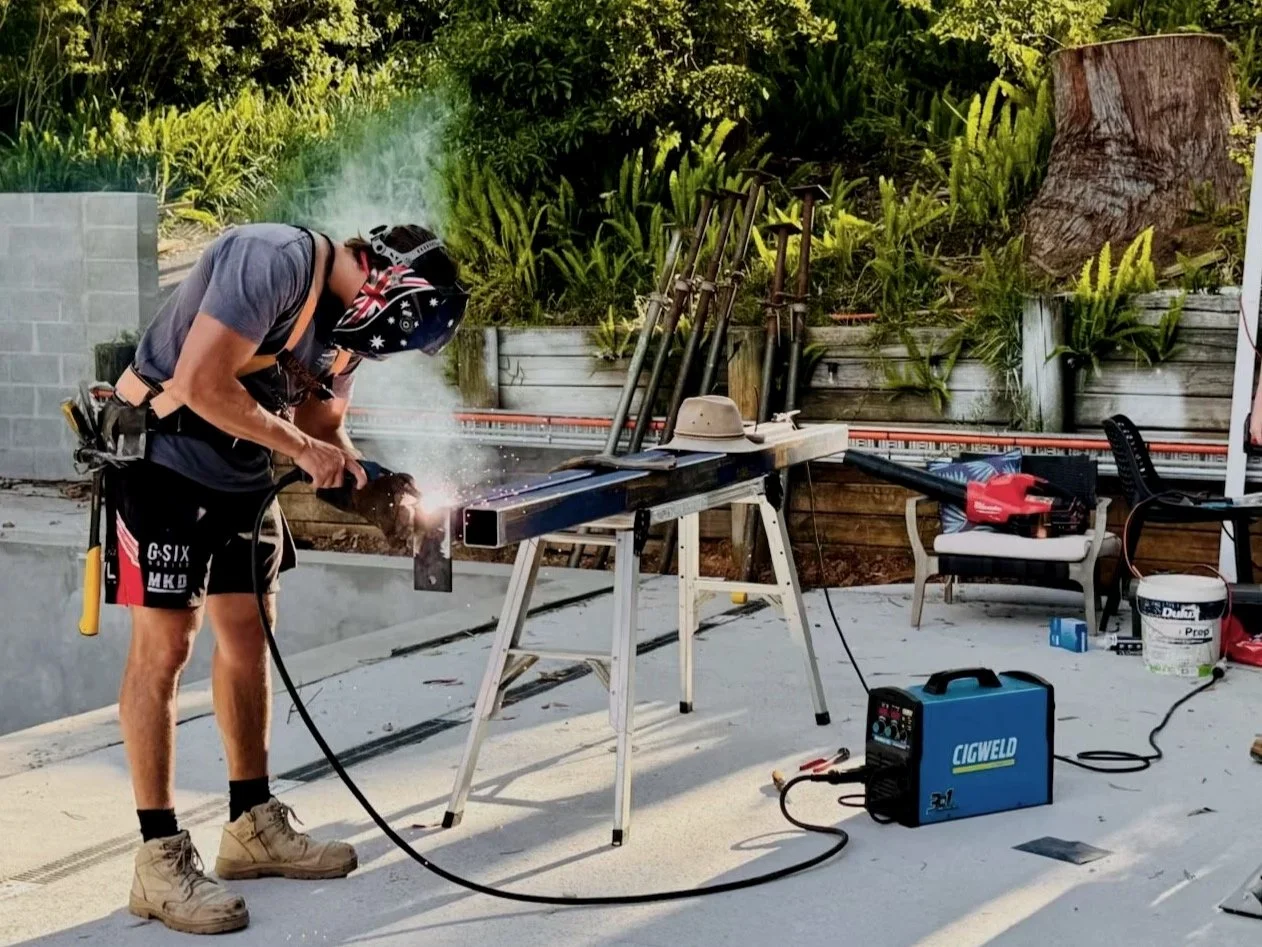 Person welding metal beam outdoors, wearing safety gear including helmet and gloves, with sparks flying, work tools and equipment nearby, plants and a tree stump in the background.