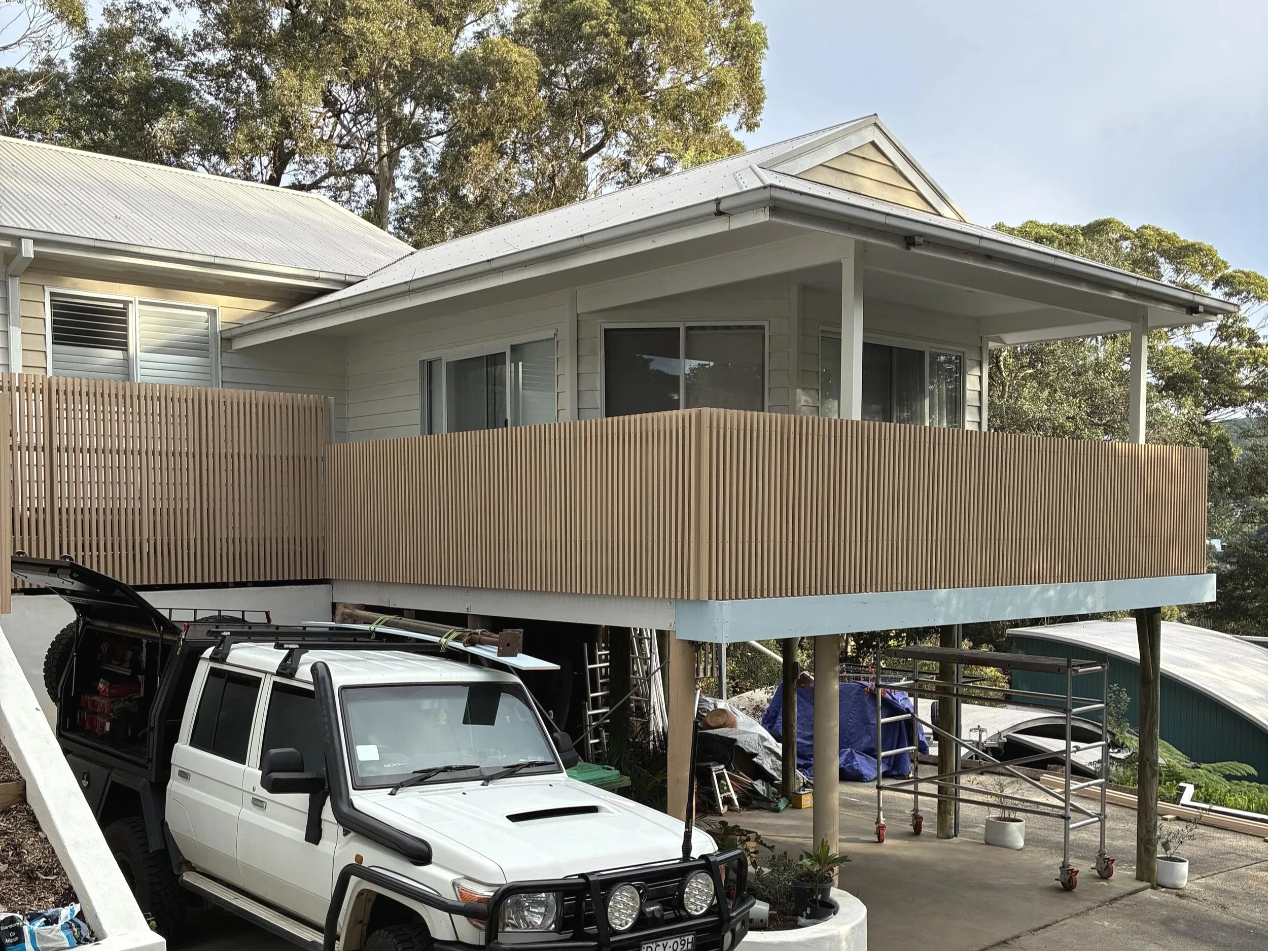 A house with a metal roof surrounded by trees, with a wooden fence in the foreground. There is a raised area under construction with building materials and tools. A white Jeep is parked in front.