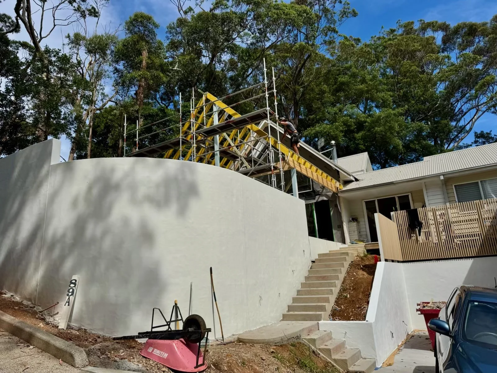 Construction workers installing a roof on a house, surrounded by scaffolding, with a white exterior wall and a staircase leading up to the entrance, in a wooded area under a blue sky.