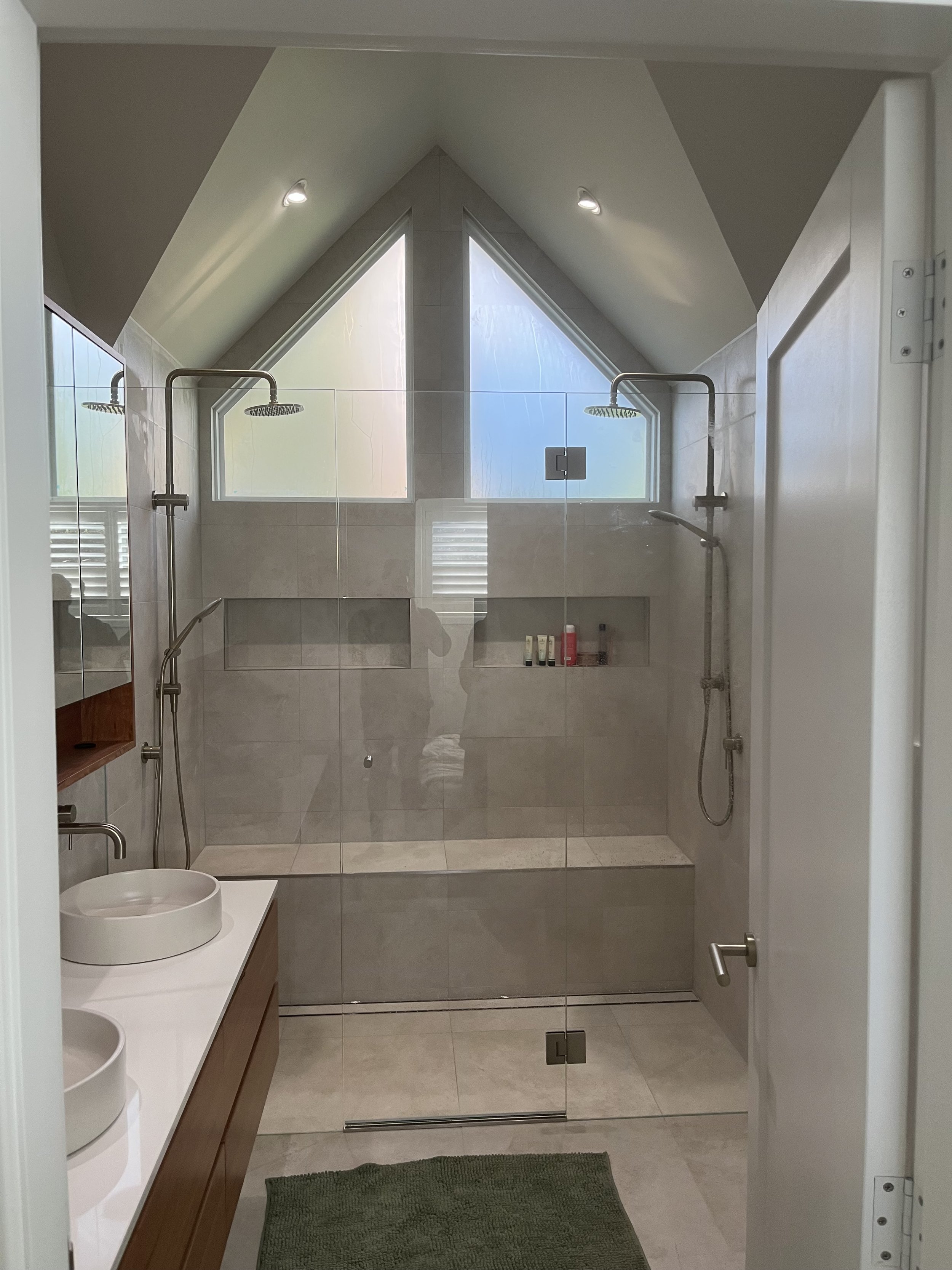 A modern bathroom with a glass shower enclosure featuring two rainfall showerheads, a built-in tiled shelf with toiletries, a white vessel sink on a wooden vanity, and a green bath mat on the tiled floor. Two triangular windows are above the shower, and there is a white door to the right.